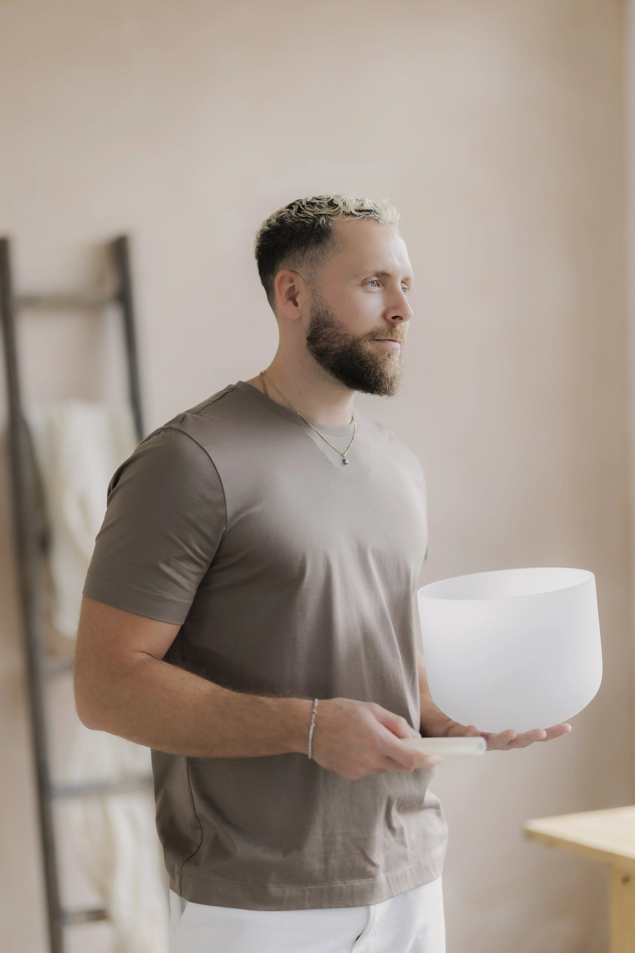 A man with a beard and short, curly hair holds a singing bowl and stands in a softly lit room with a beige wall and a blurred ladder in the background.