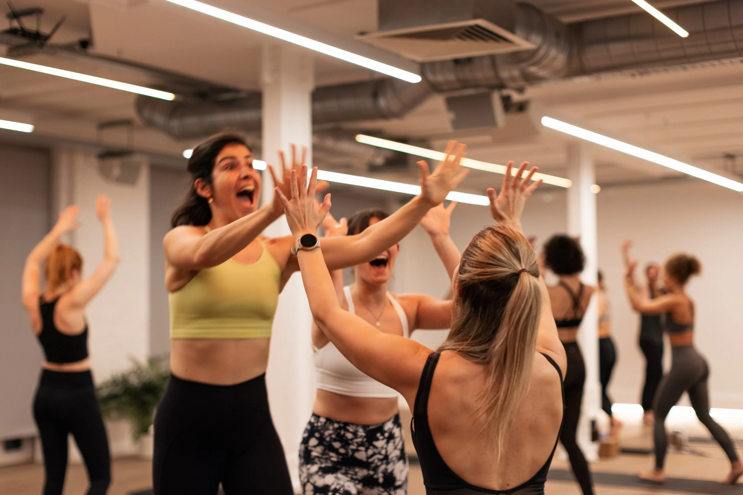 Women participating in a yoga class, reaching towards each other in a brightly lit, modern studio with high ceilings and exposed ductwork.