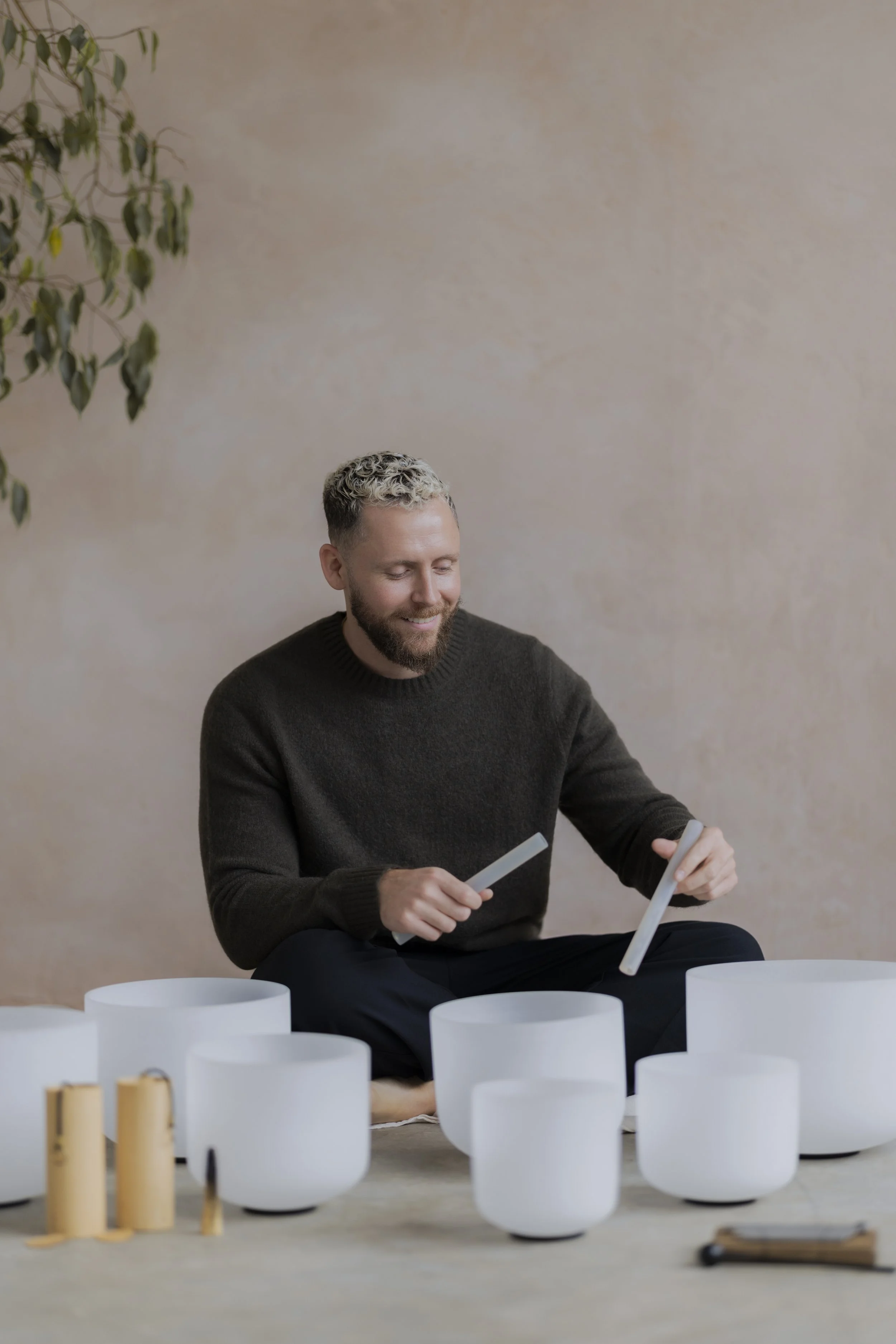A man with a beard and curly blonde hair sitting on the floor, playing crystal singing bowls, with a neutral beige wall and a plant in the background.