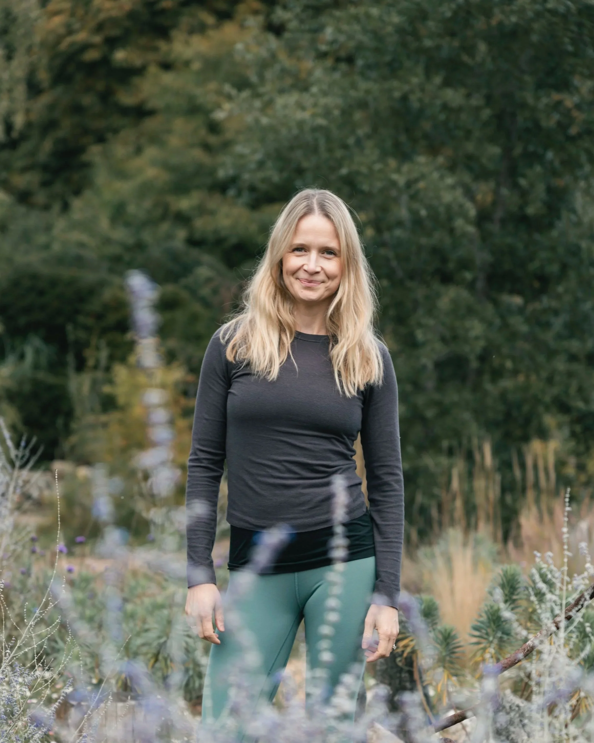 A woman with long blonde hair, wearing a black long-sleeve shirt and teal pants, standing outdoors among greenery and purple flowers, smiling at the camera.