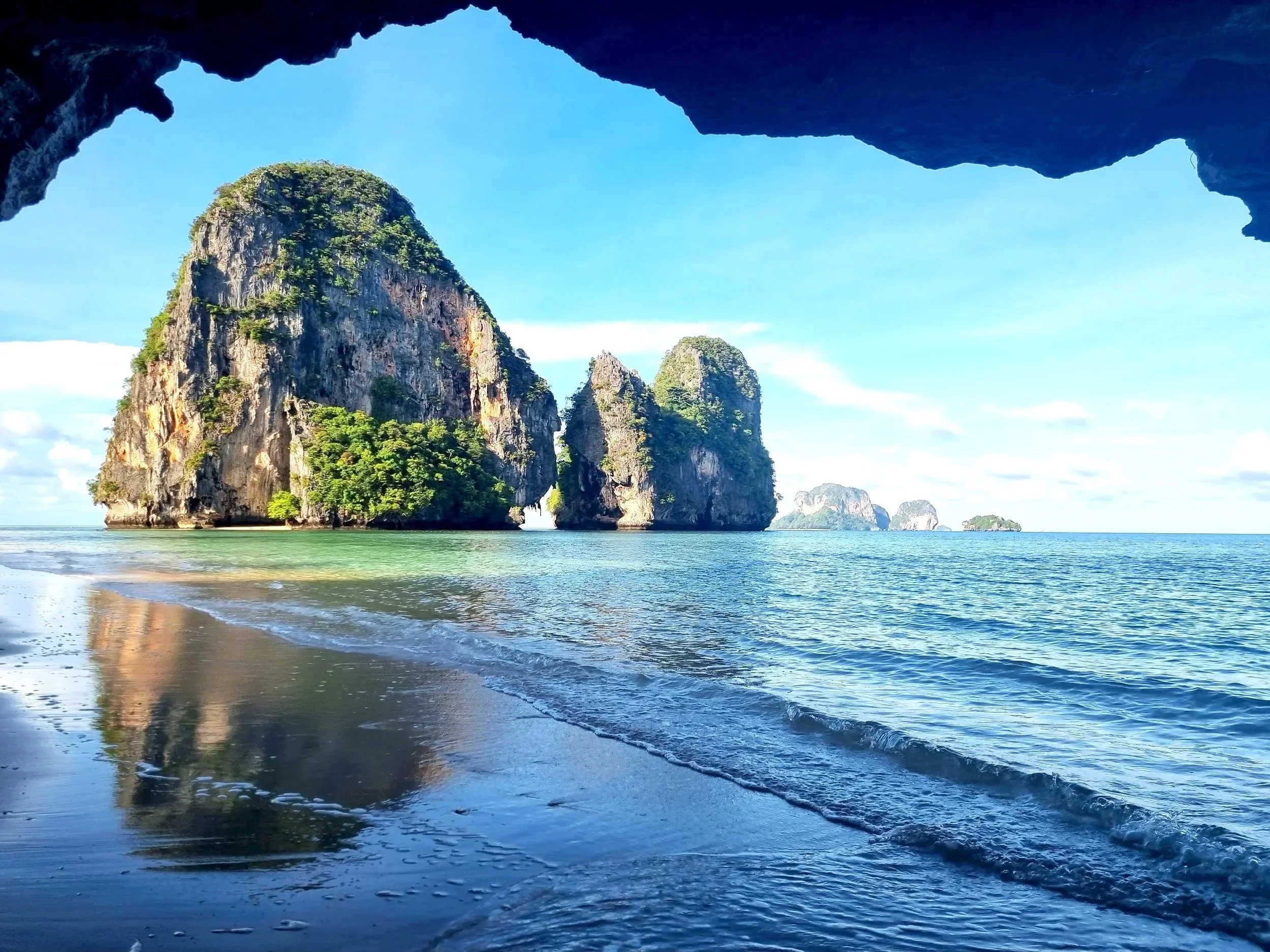 View from inside a cave looking out to a tropical beach with large limestone islands in the distance, clear blue water, and a partly cloudy sky.