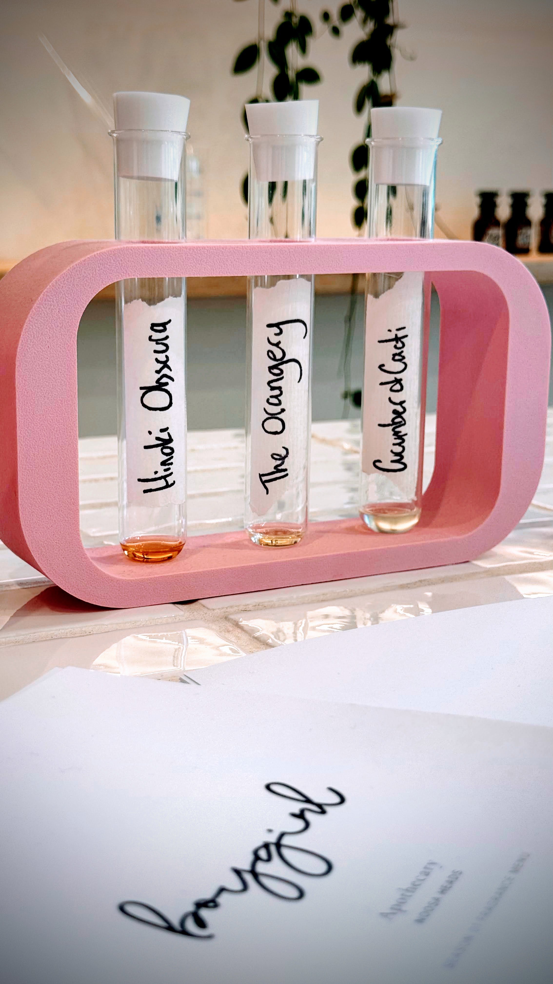 Three test tubes labeled 'Hindu Oshucha', 'The Ocaragy', and 'Gundachi' are in a pink holder. They contain handmade perfumes mixed on-site at the salon.