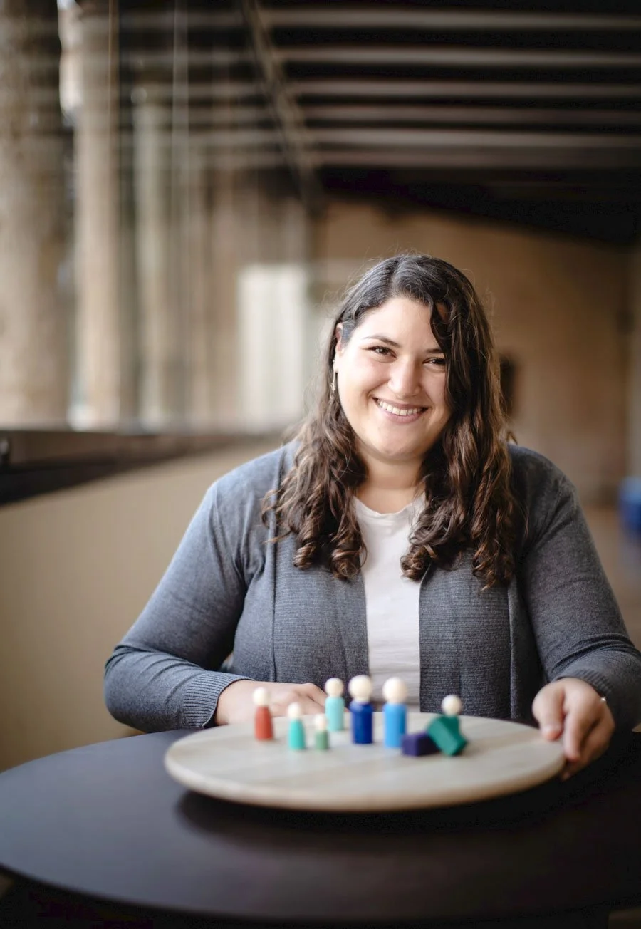 A woman with long, wavy brown hair smiling and sitting at a table with a wooden board displaying small, colorful game pieces.
