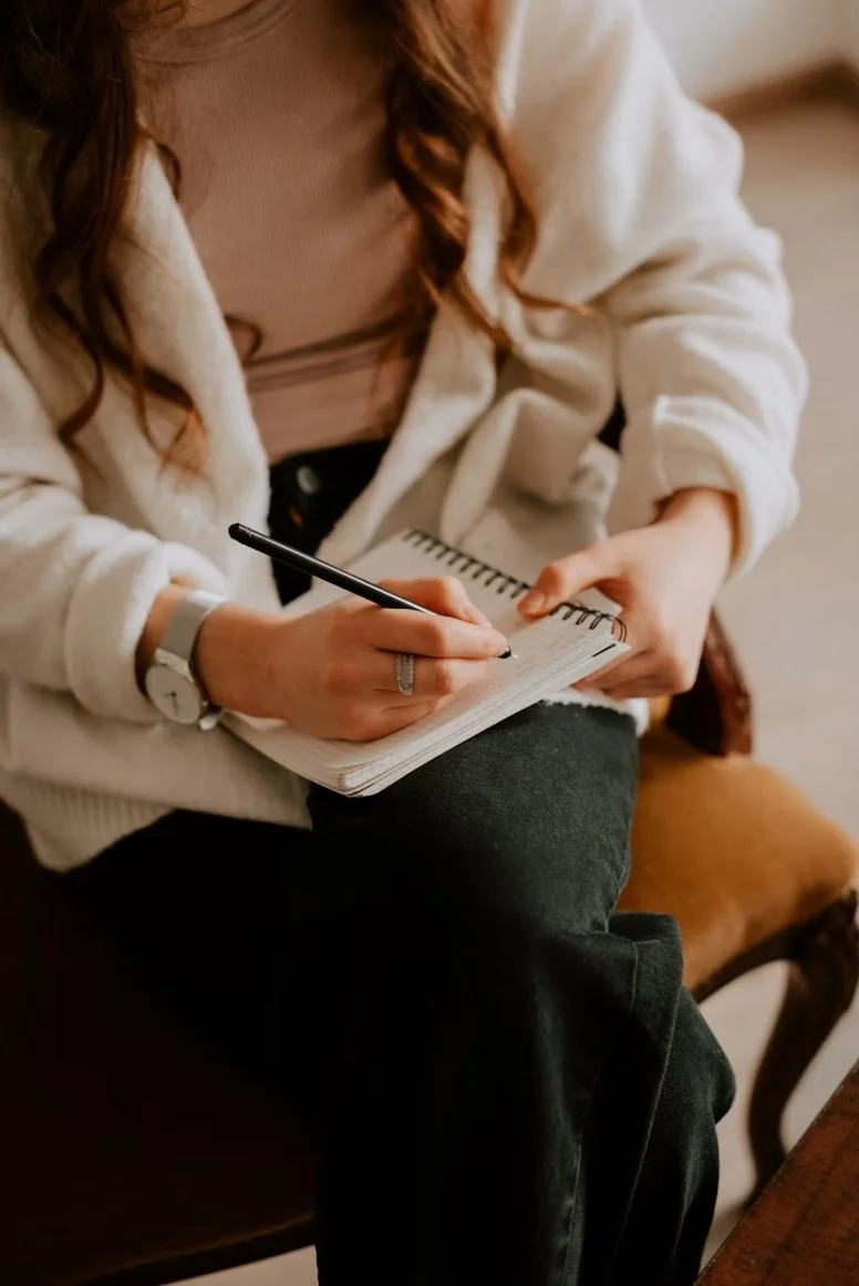 A woman with long, wavy hair wearing a white sweater and a beige top, sitting on a chair, writing in a spiral notebook with a black pen.