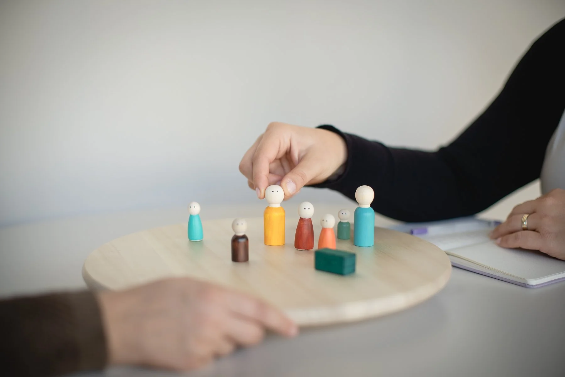 A person arranging small wooden figurines on a round wooden table, with a notebook and pen nearby.