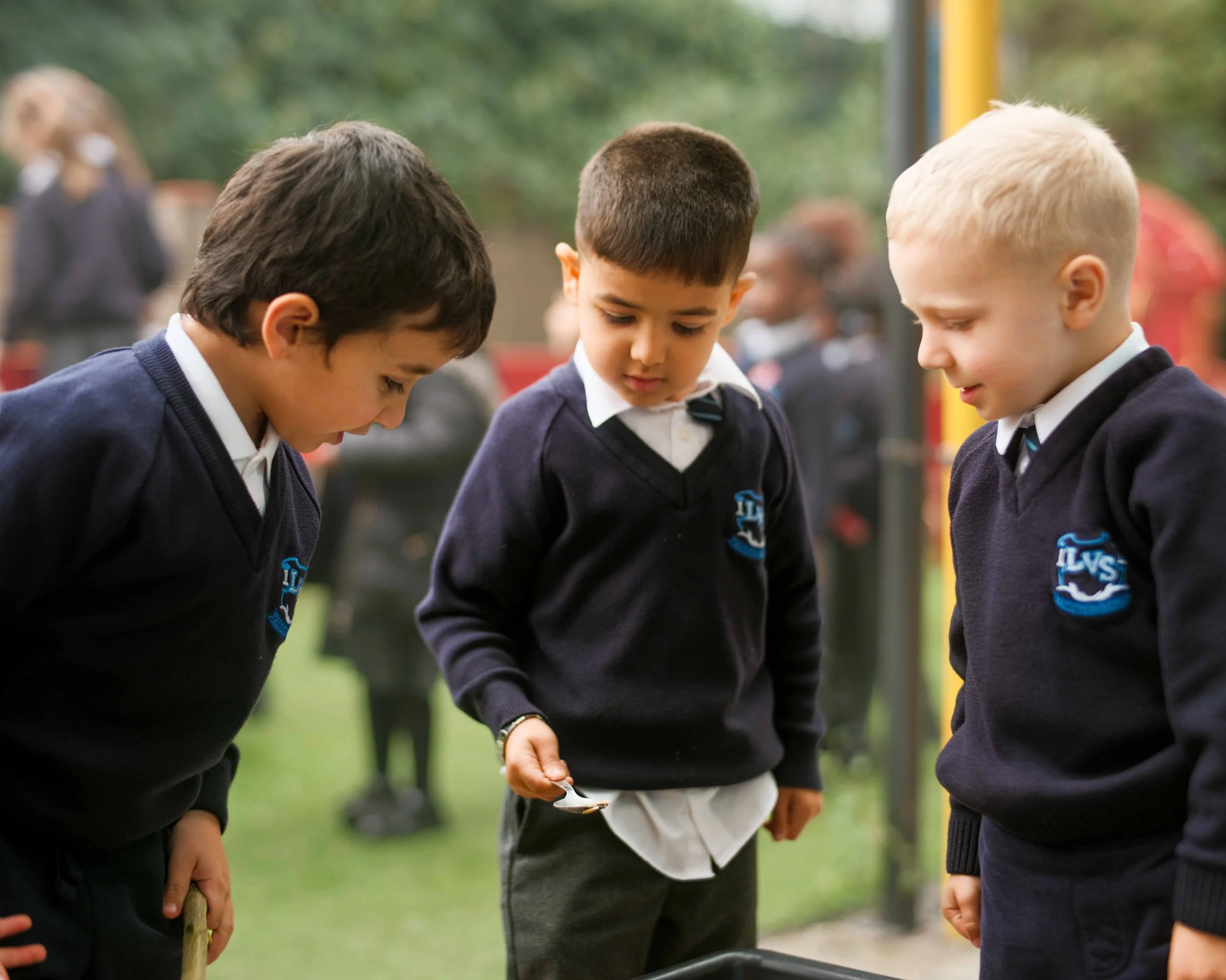 Three young boys in school uniforms are looking down at something, outside on a grassy area with blurred trees and other children in the background.