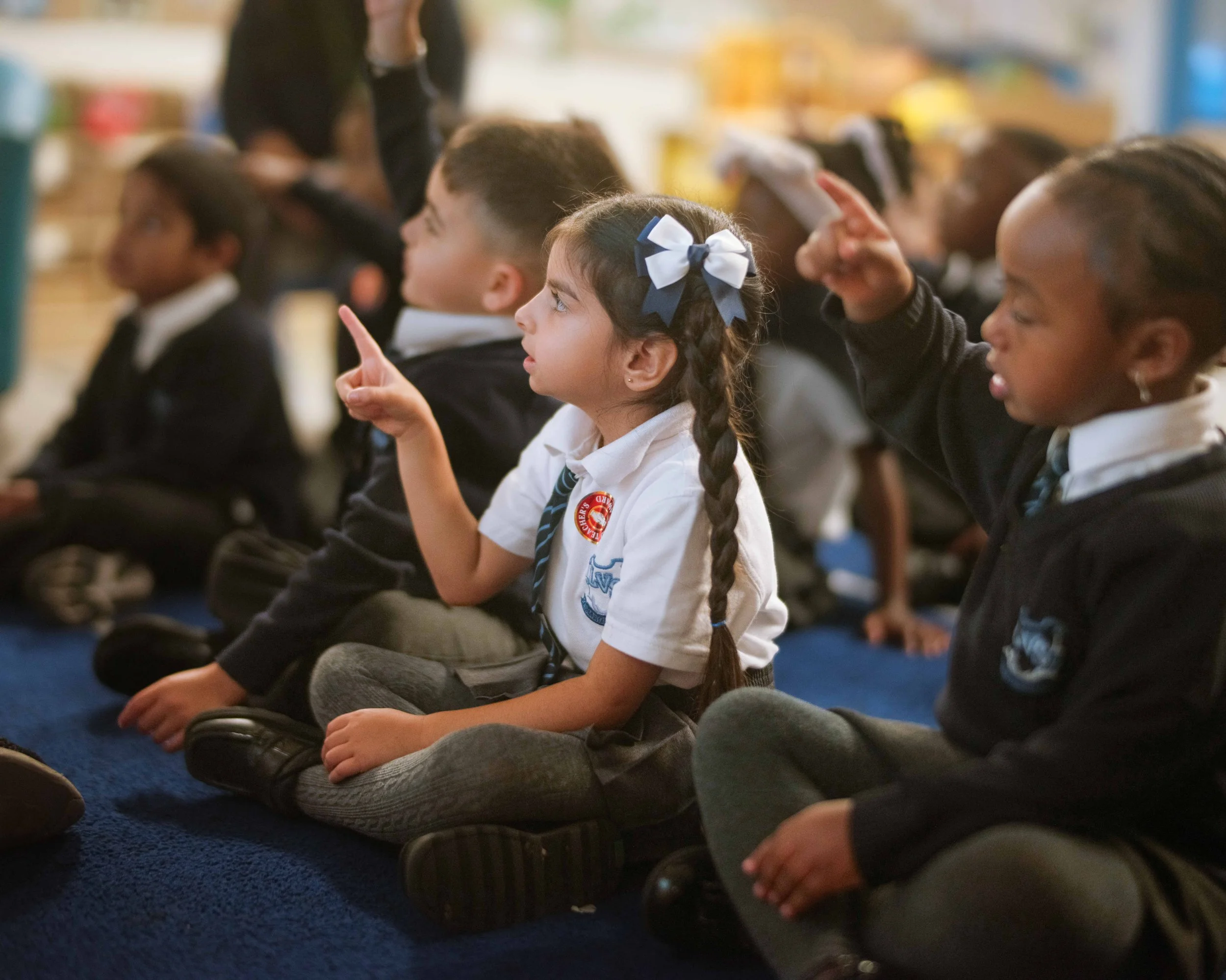 A group of young children sitting cross-legged on a blue carpet during a school activity. One girl with long braids and a navy and white ribbon in her hair is pointing upward, engaged in the lesson.