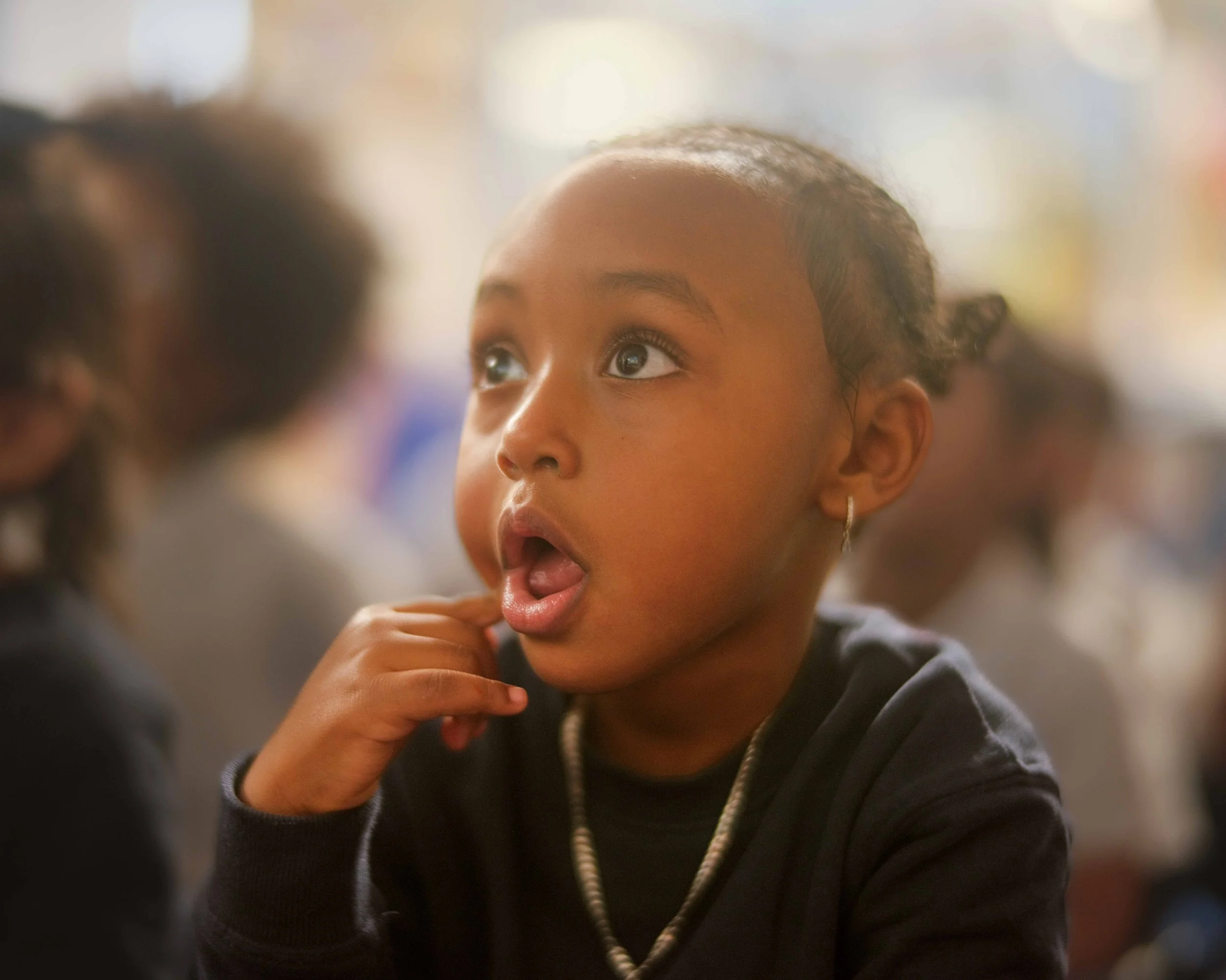 Close-up of a young girl with curly hair and hoop earrings, looking upward, with a surprised or curious expression. She is wearing a dark hoodie and a beaded necklace, with a blurred background.
