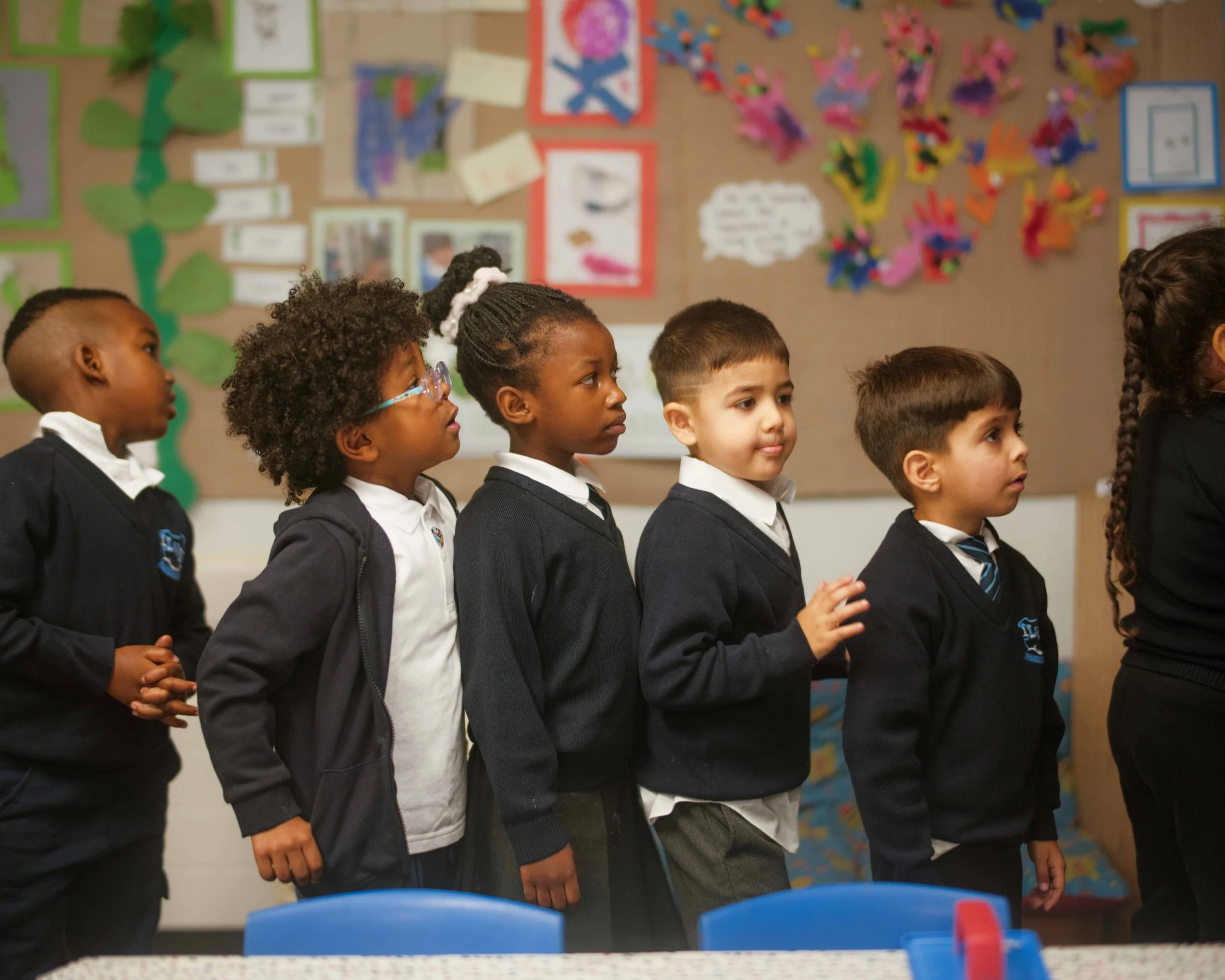 A group of children standing in a line in a classroom, paying attention to something out of the frame. The children are wearing school uniforms and are of varied ethnic backgrounds. The background shows colorful wall decorations and educational posters.
