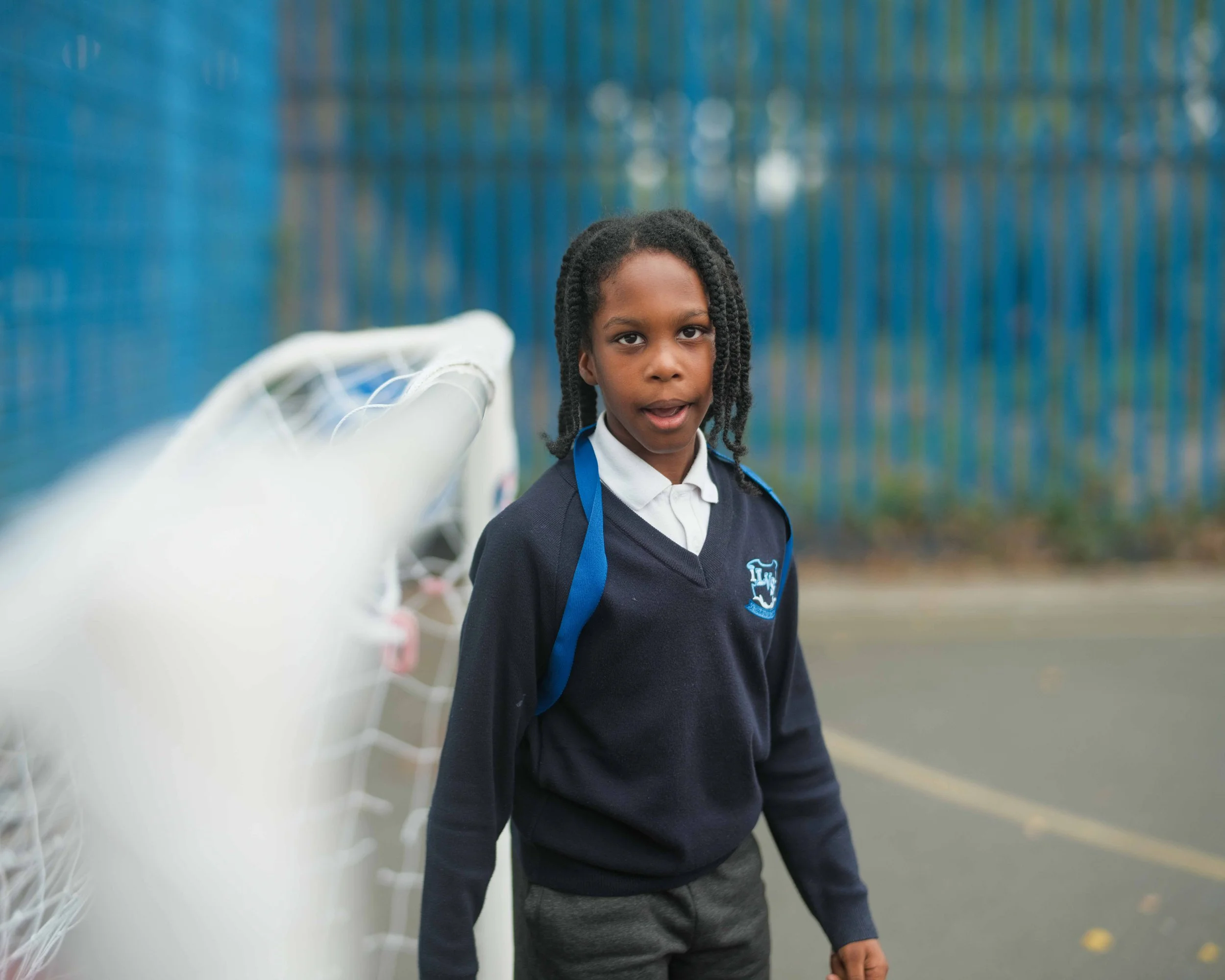 A young girl with braided hair wearing a navy blue school uniform with a white shirt and blue tie, standing outdoors near a tennis court, with a blue fence in the background.