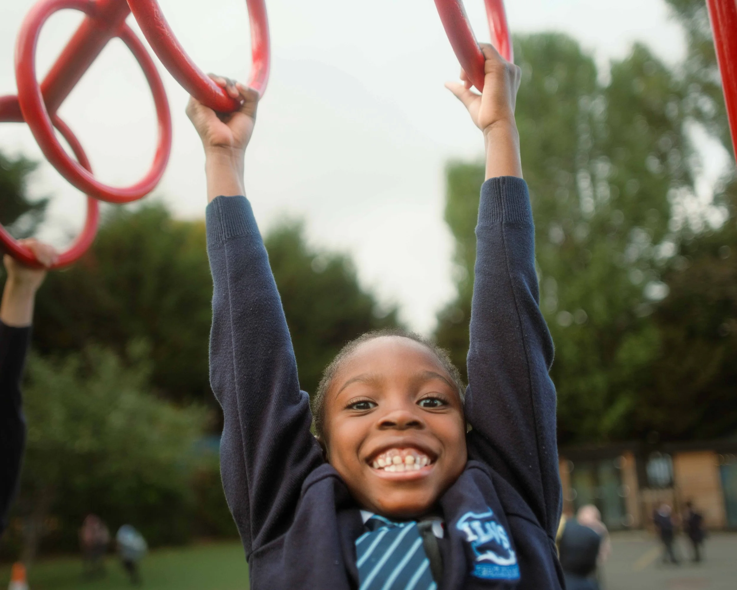 Smiling child hanging from red playground equipment outdoors with blurred trees in background.