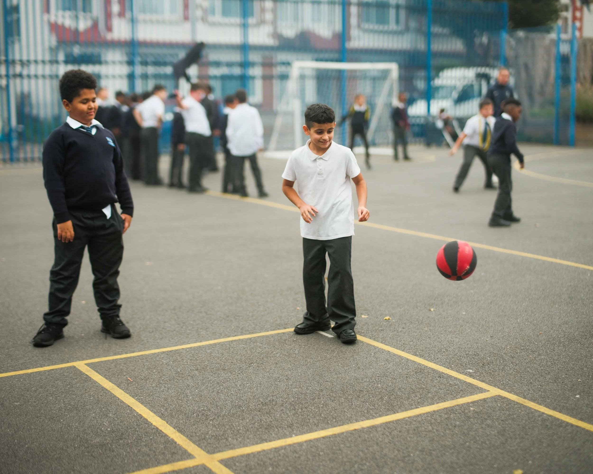 Young boys playing with a red and black ball on a school playground, with students and a teacher in the background.
