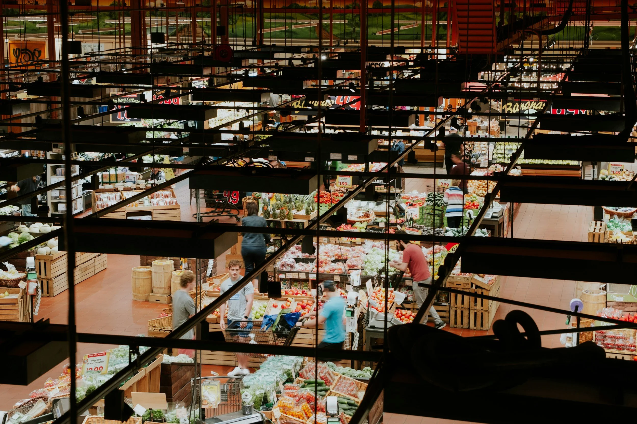 View of a grocery store's produce section with customers shopping for fruits and vegetables, seen through the store's ceiling framework.