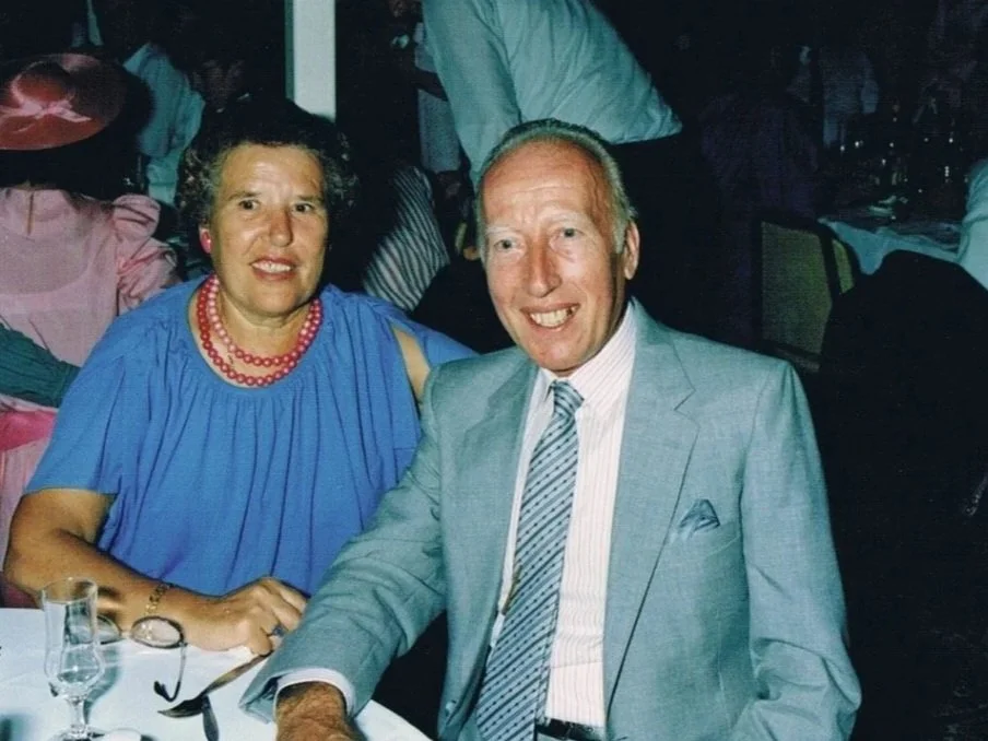 A smiling woman in a blue dress with a red necklace sitting next to a man in a light grey suit and striped tie at a formal event table with glasses and silverware, in a dimly lit social gathering.