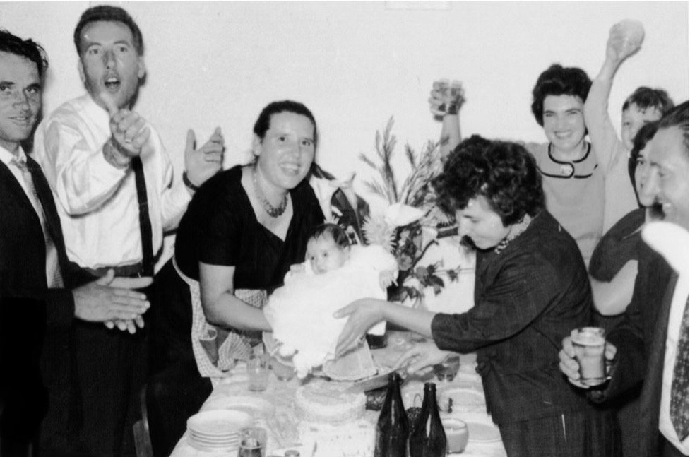 A group of people celebrating a birthday party around a table with a baby in the center, who is being handed a cake. The group appears happy, with some holding drinks and others smiling or raising their hands in celebration.