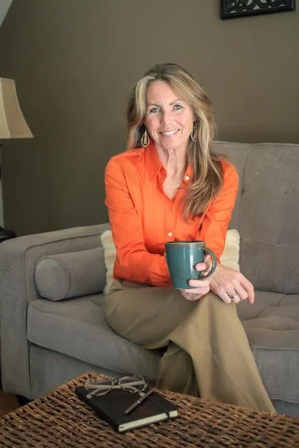A woman with blonde hair is sitting on a beige couch, smiling, and holding a teal coffee mug. She is wearing an orange shirt and beige pants in a cozy living room.