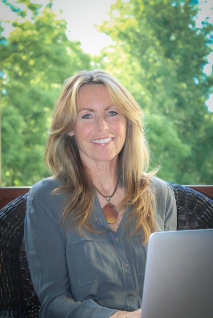 A woman with long blonde hair sitting outdoors with a laptop, green trees in the background, smiling at the camera.