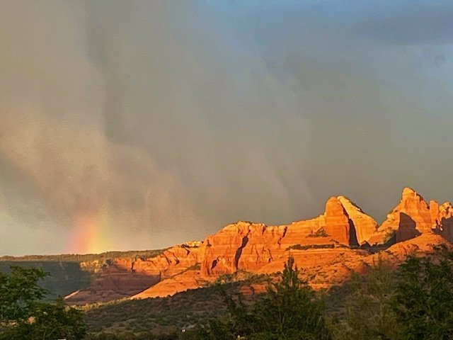 Sunset over red rock formations with a rainbow in the sky and some trees in the foreground.