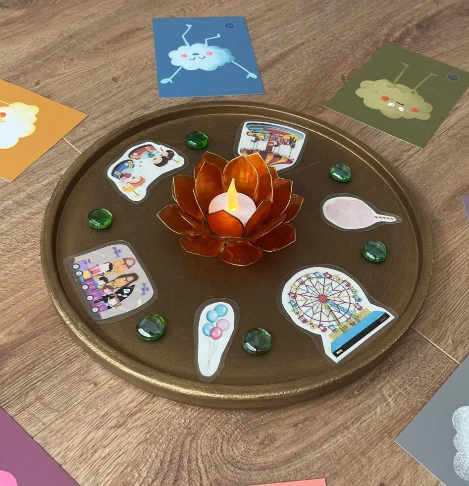 Auf einem runden goldenen Tablett in der Mitte eine dekorative Kerze in Form einer Blume. Rund um die Kerze liegen bunte Spielkarten, die verschiedene Attraktionen und Gegenstände zeigen, sowie grüne Glassteine.