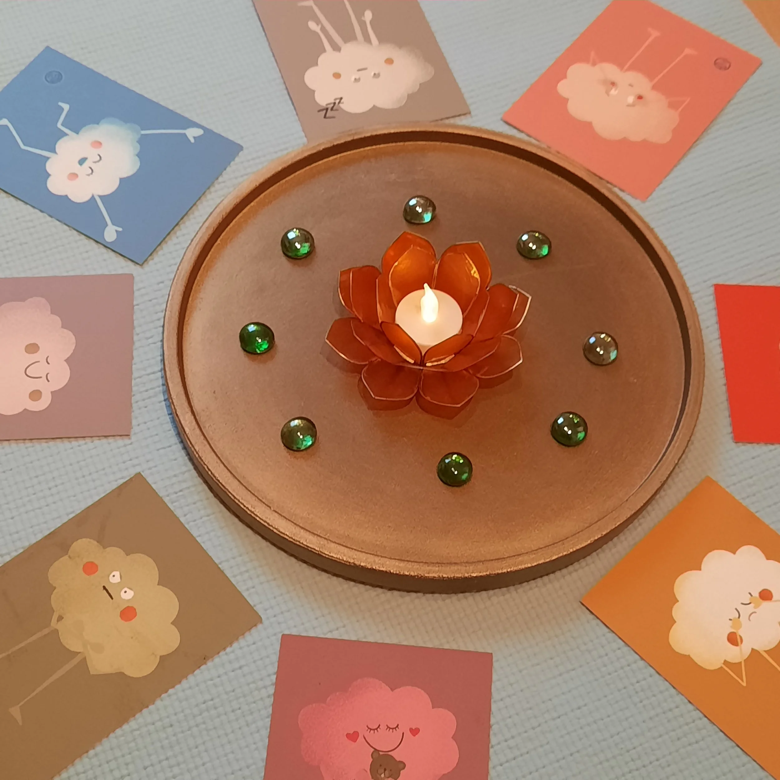 Eine Kerze in einer Blumenform in der Mitte eines runden Tellers, umgeben von bunten Karten mit Wolkenmotiven, die verschiedene Gefühle darstellen,  und kleinen grüne Dekoglassteine.