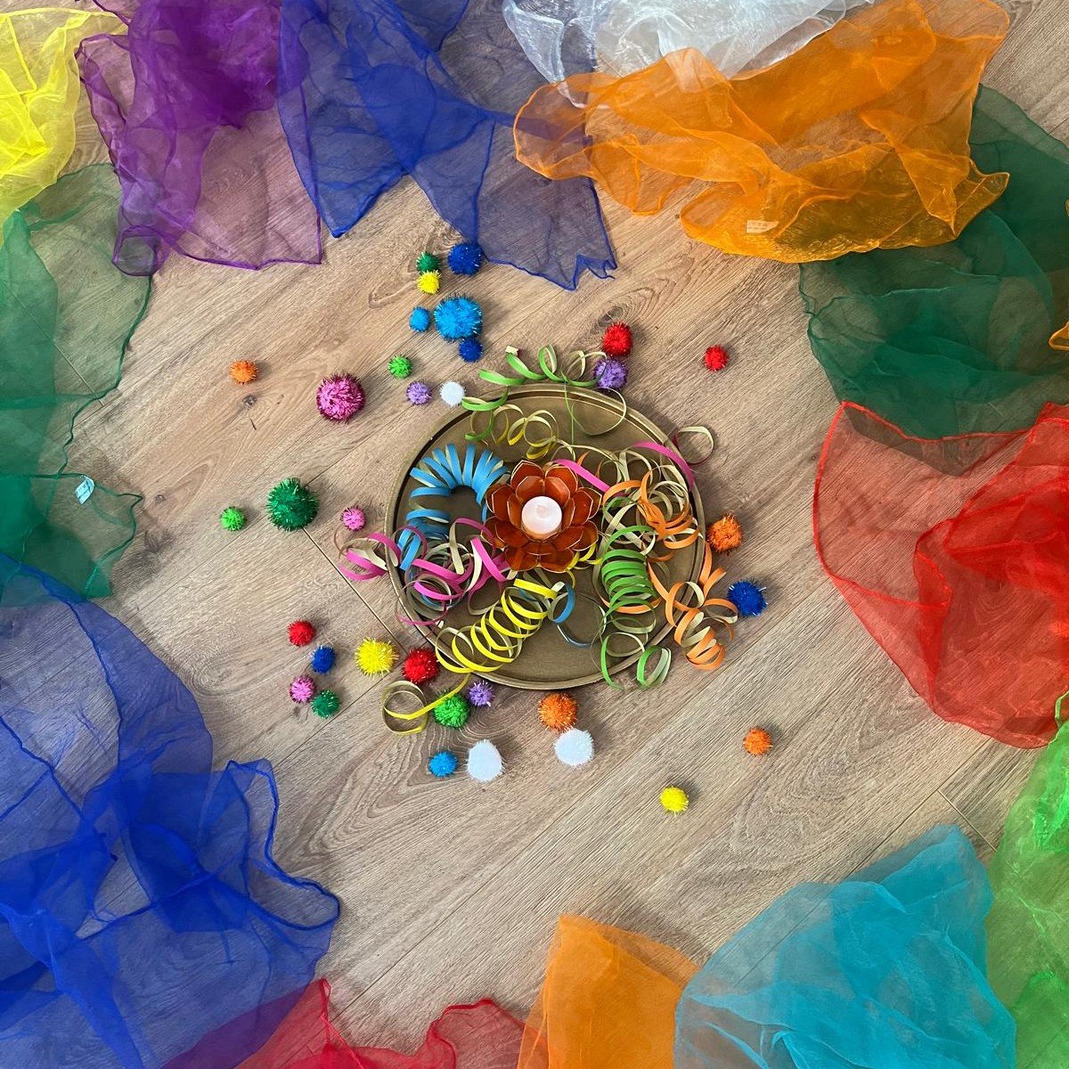 Farbenfrohe Dekoration mit bunten Luftschlangen, Pom-Poms und einer Kerze in der Mitte, umgeben von Chiffontüchern in Regenbogenfarben auf einem Holzboden.