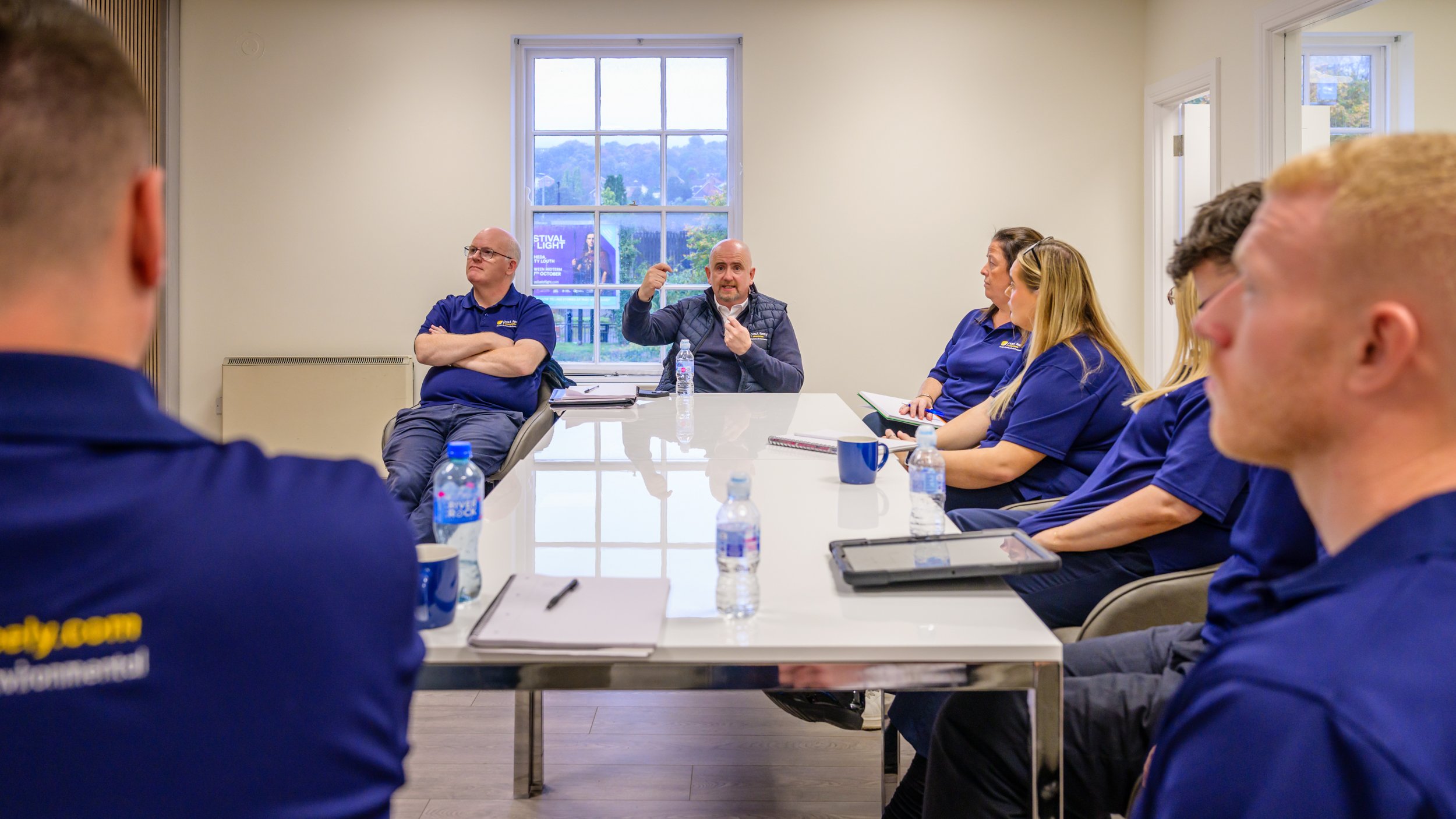A group of people in blue shirts sitting around a conference table during a meeting, with a man in the center speaking and gesturing with his hand.