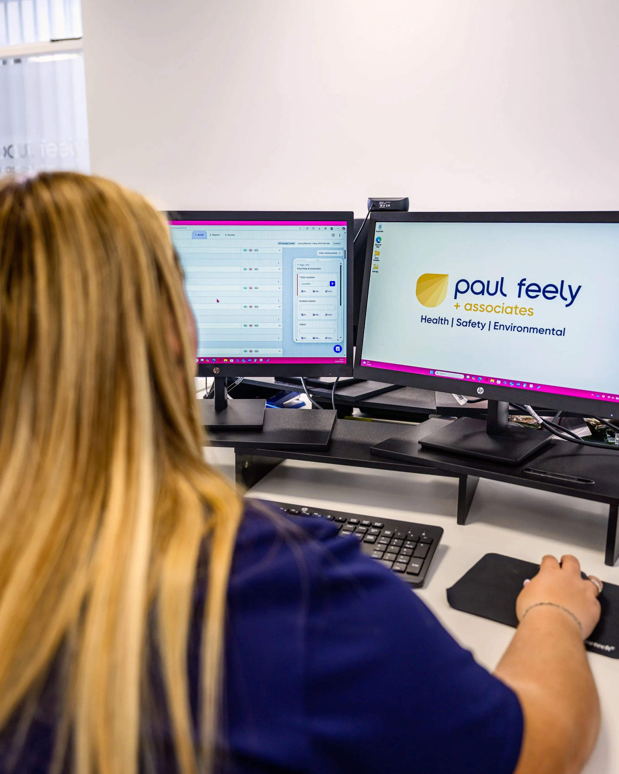 A woman working at a desk with dual computer monitors, one displaying a scheduling application and the other showing a presentation slide for Paul Feely + Associates, focusing on health, safety, and environmental services.
