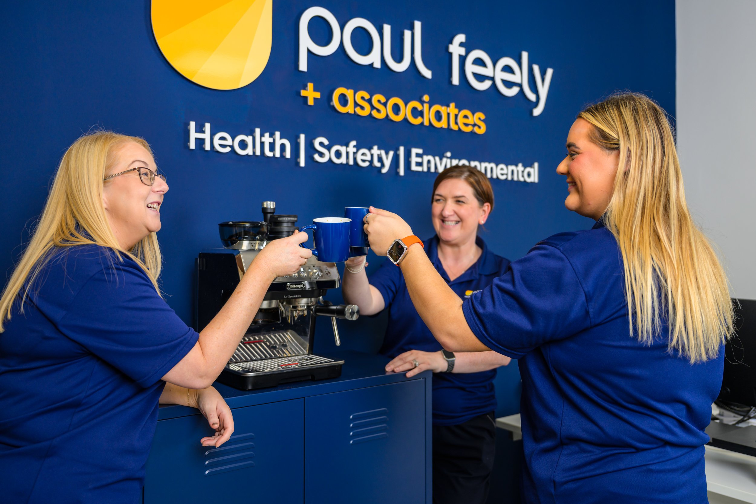 Three women in matching blue shirts sharing a laugh and clinking coffee cups behind a coffee machine at Paul Feely + Associates office, with blue wall displaying company's name and services.