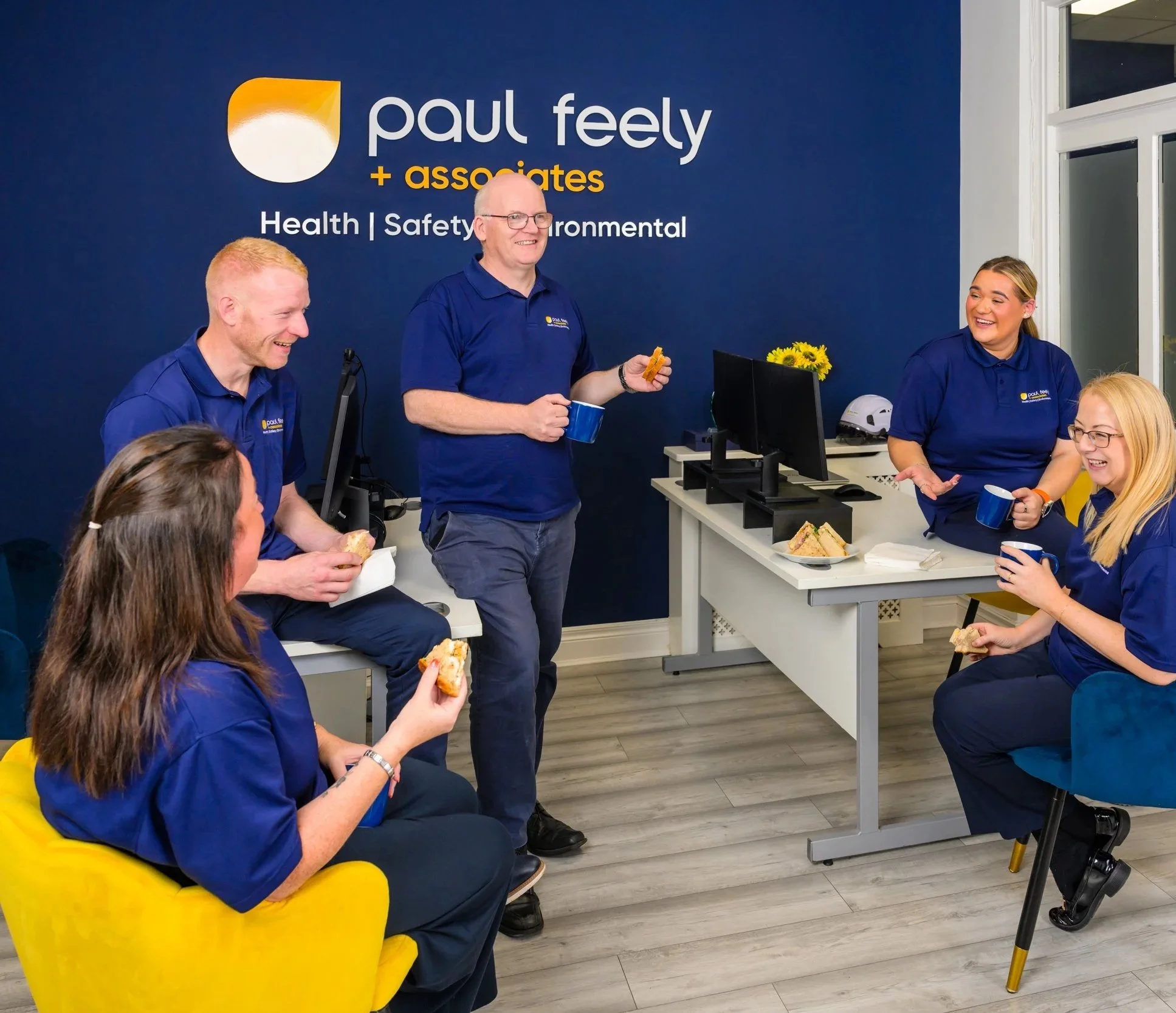 Group of six people in blue uniforms enjoying sandwiches and coffee in an office, with a dark blue wall with the logo 'Paul Feely + associates' and the words 'Health | Safety | Environmental'.