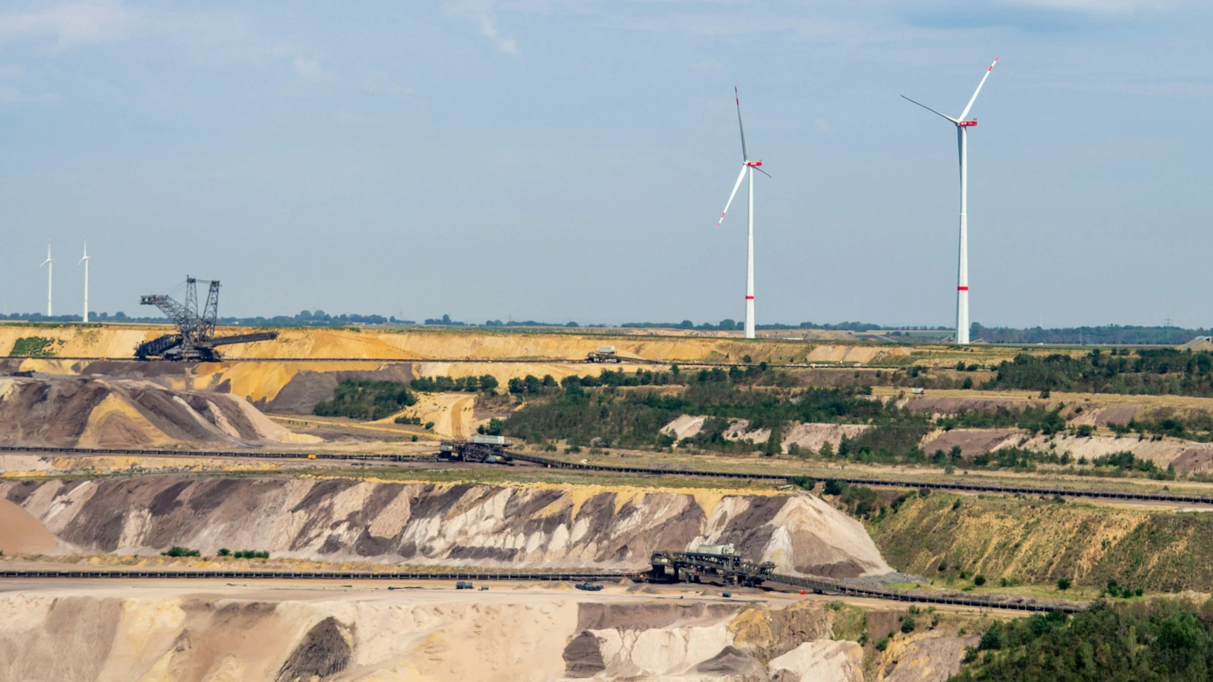 View of a large industrial or mining site with multiple wind turbines in the background, a construction crane, and several trucks transporting materials across the terrain under a blue sky.