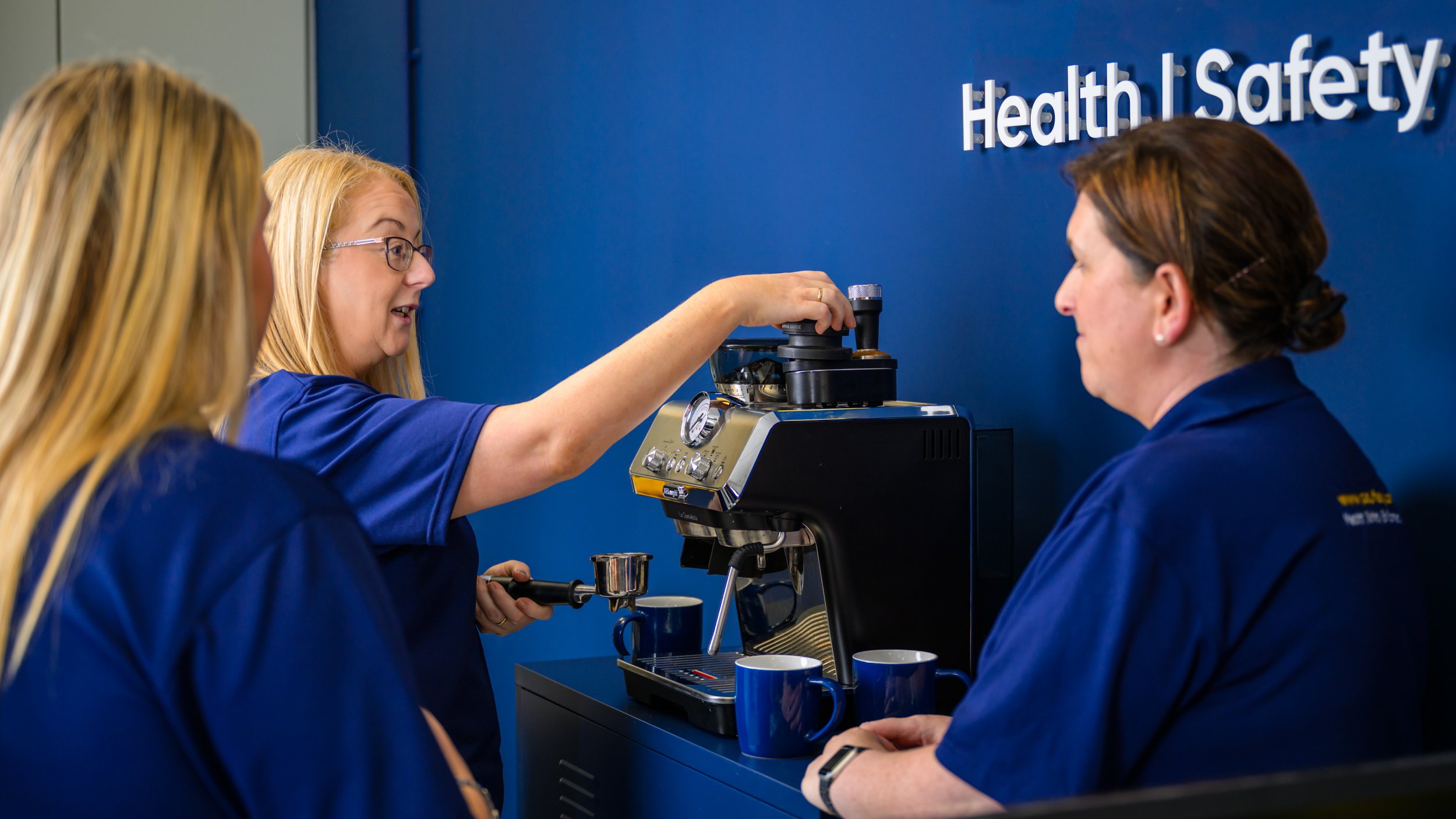 Three women in blue scrubs making coffee with an espresso machine, with a blue wall background and the words 'Health & Safety' on the wall.