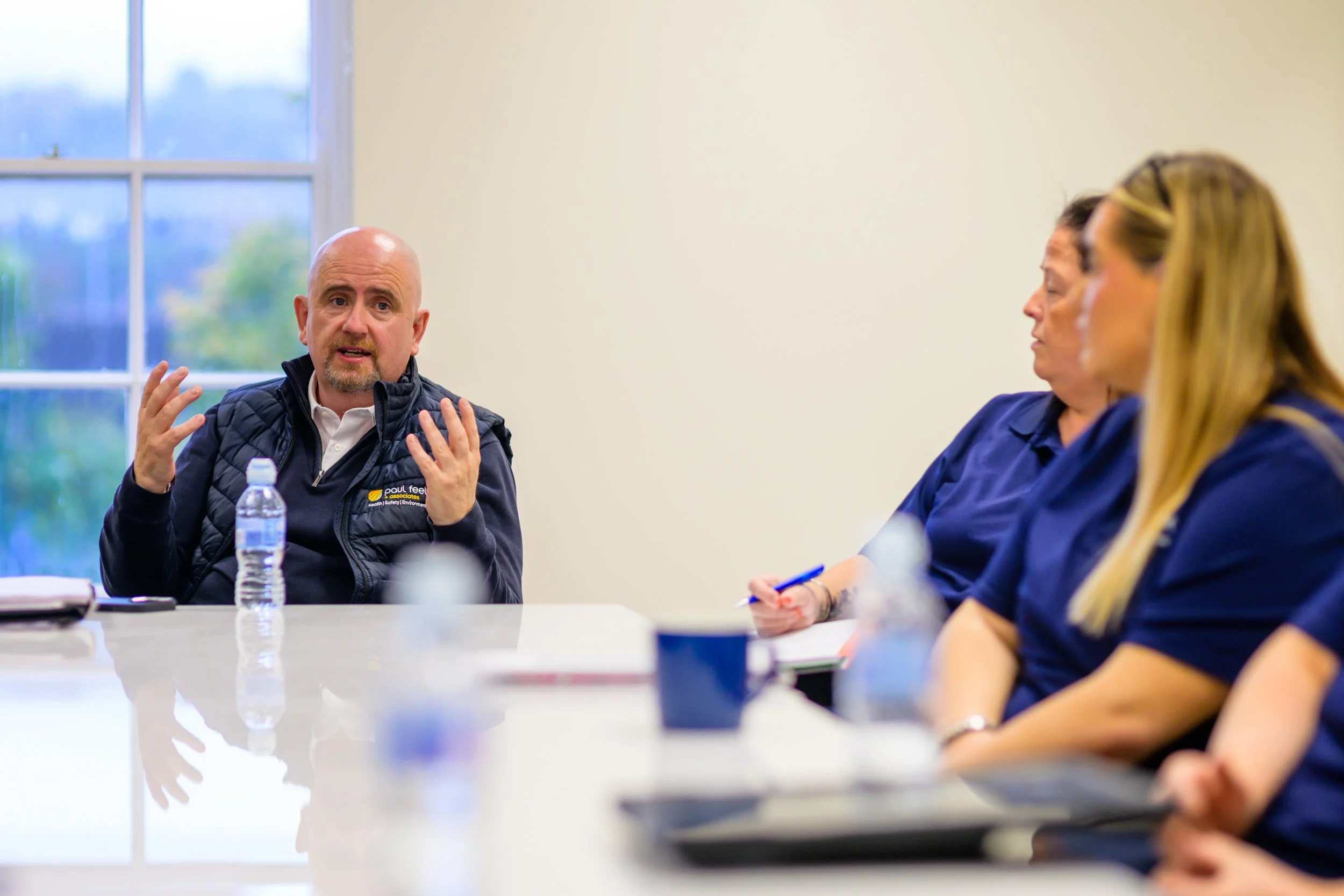 A man speaking during a meeting, with two women listening, in a room with a window showing a blurred outdoor scene.