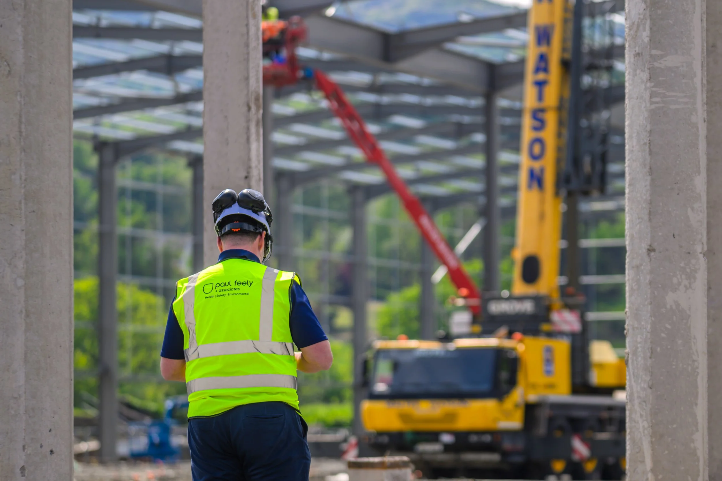 A construction worker wearing a yellow vest and a safety helmet looks at a large yellow crane with a red arm at a construction site.