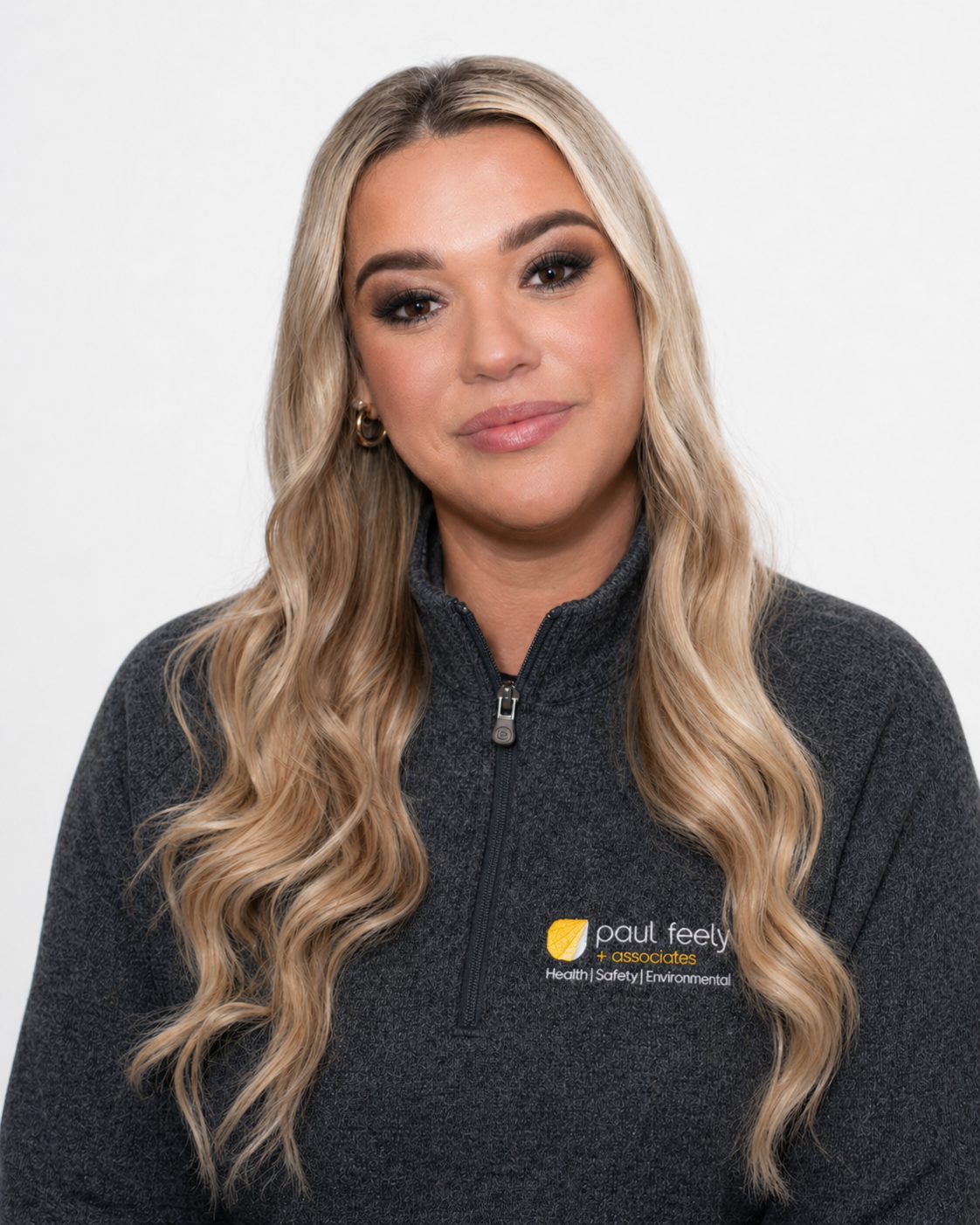 A woman with long, wavy blonde hair wearing a dark gray zip-up jacket with a logo that reads 'pault feely + associates' and mentions health, safety, and environmental.