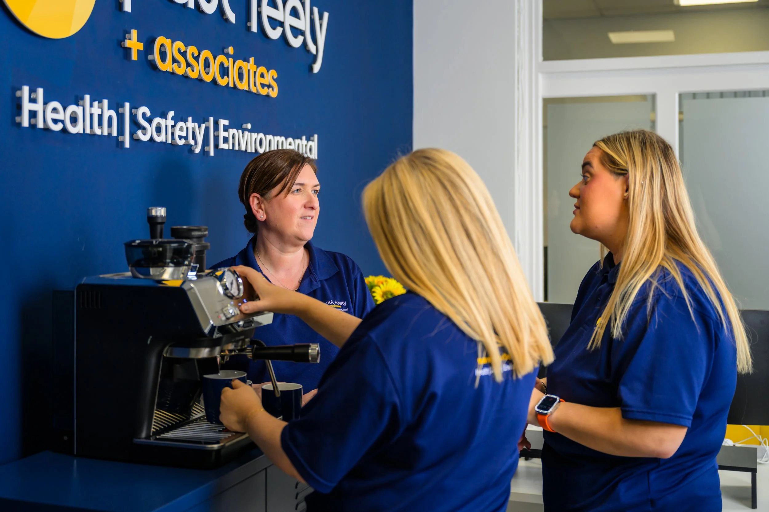 Three women in matching blue shirts in a workplace break room or office with a blue wall displaying the name 'Healthly + associates' and words like 'Health', 'Safety', 'Environmental'. One woman is operating a coffee machine while the other two women