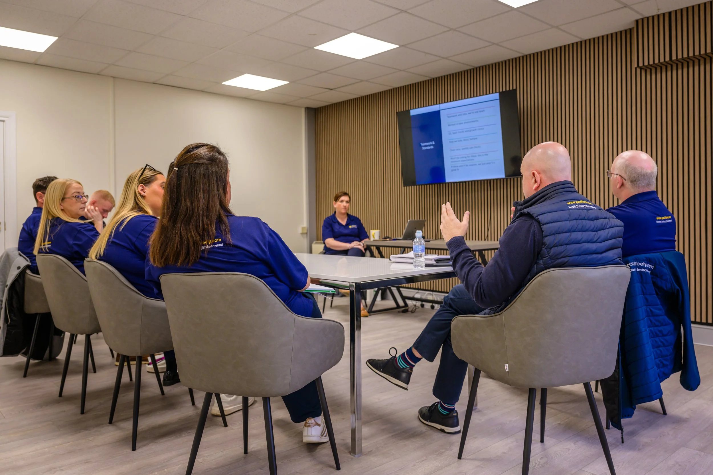 A group of people attending a meeting or training in a modern conference room with a large screen displaying a presentation.
