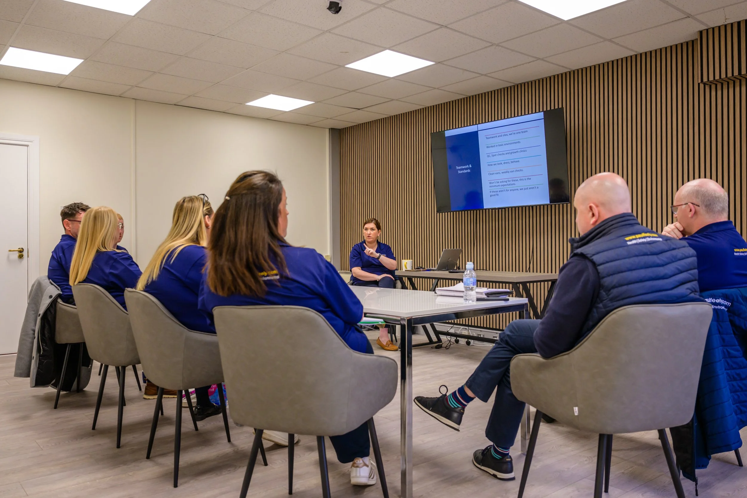 People in a meeting room listening to a woman presenting. The room has a wood-paneled wall and a large screen displaying a presentation slide titled 'Teamwork & Standards.'
