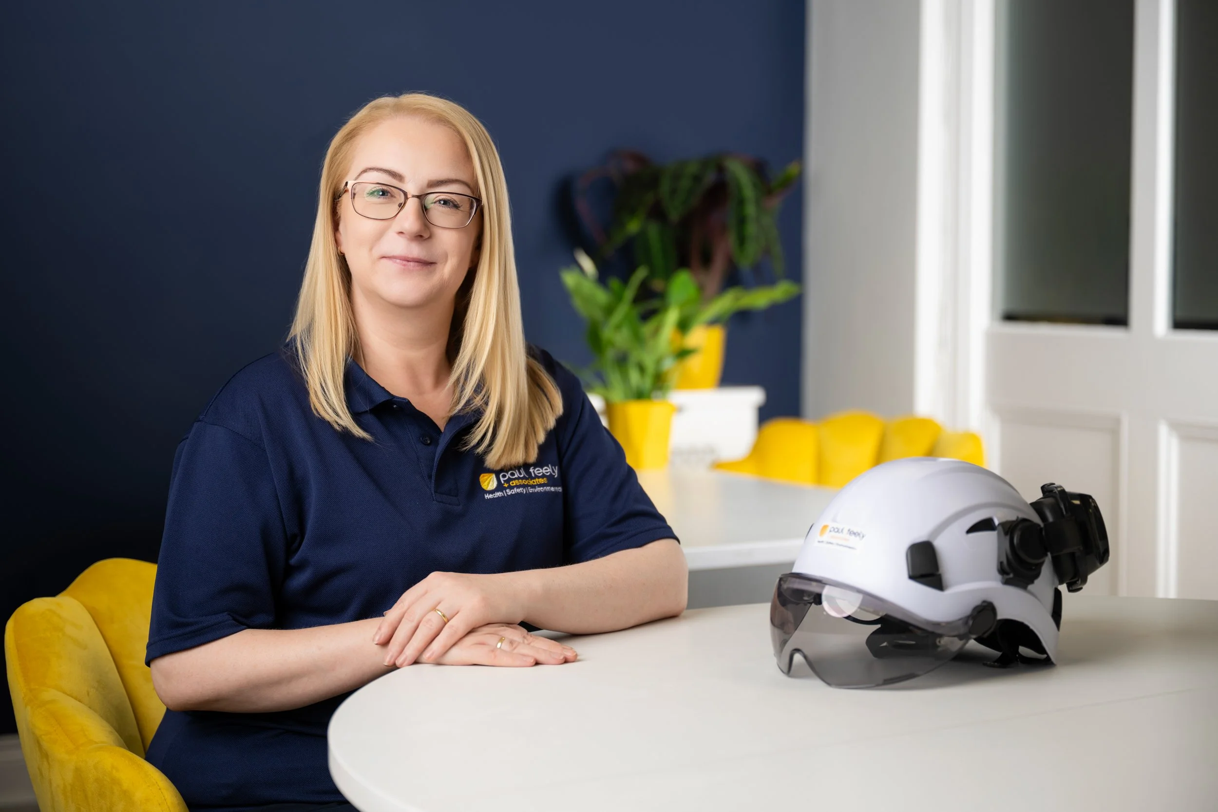 A woman with blonde hair and glasses sitting at a white table with a white safety helmet and a virtual reality headset in front of her in a modern office with yellow chairs and a plant in the background.