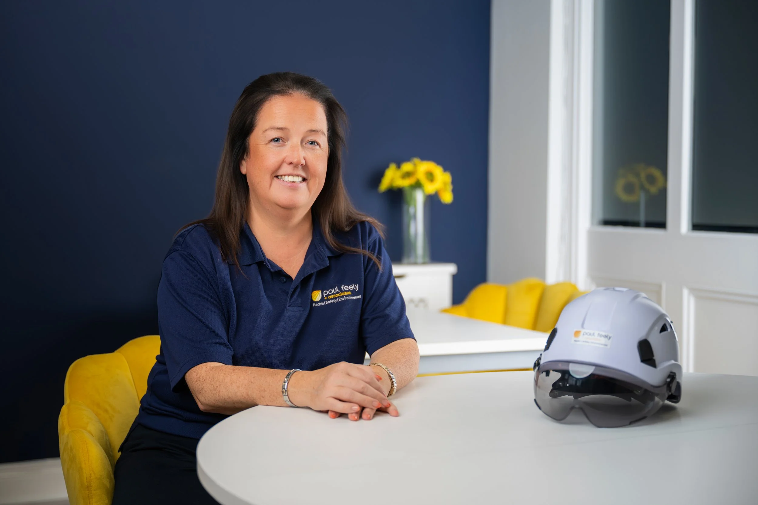 A woman with long dark hair wearing a navy blue shirt is seated at a white table in a room with yellow chairs and a dark blue wall. She is smiling and looking at the camera. On the table, there is a white helmet with black accents and a yellow logo. In the background, there is a vase with yellow flowers on a white side table and windows with white frames.