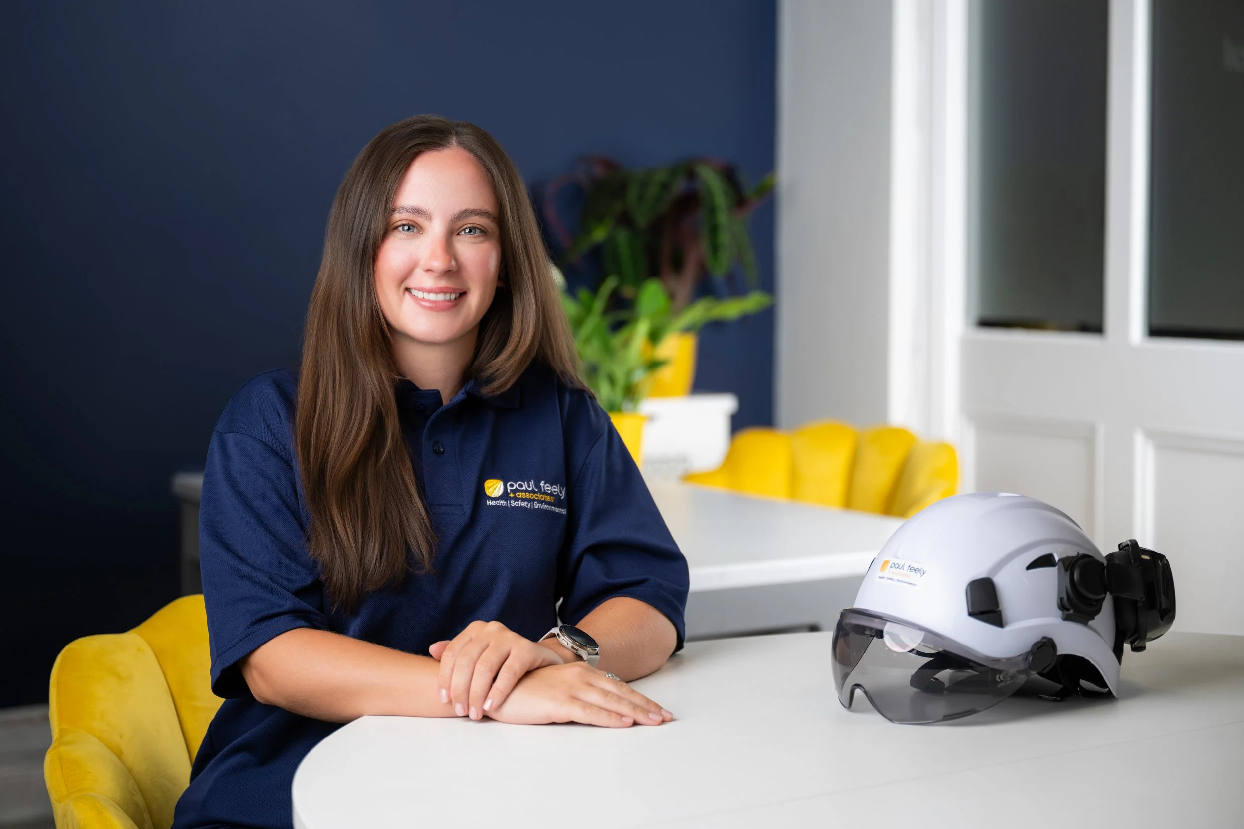 A woman with long brown hair wearing a blue polo shirt sitting at a white table with a white helmet and a virtual reality headset on the table. She is smiling and sitting in a bright, modern room with a yellow chair, green plants, and a dark blue wall.