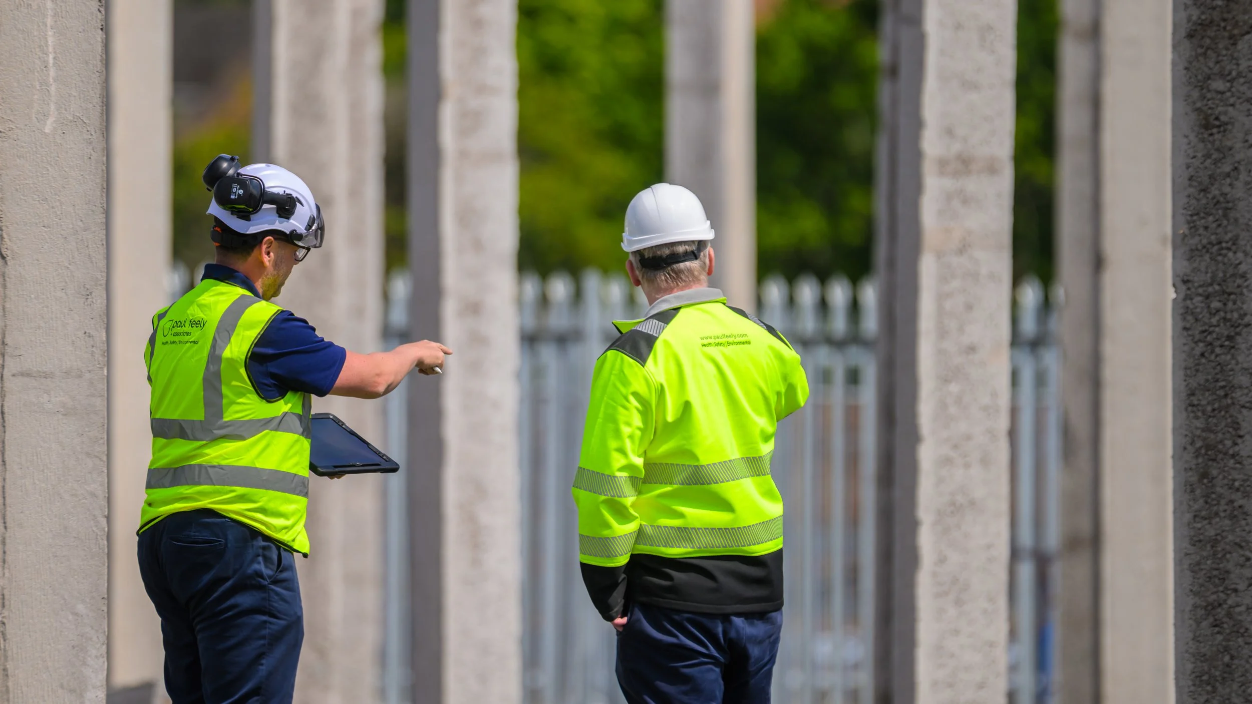 Two construction workers in high-visibility jackets and white safety helmets stand among large concrete pillars outdoors; one worker points at something while holding a tablet, and the other listens.