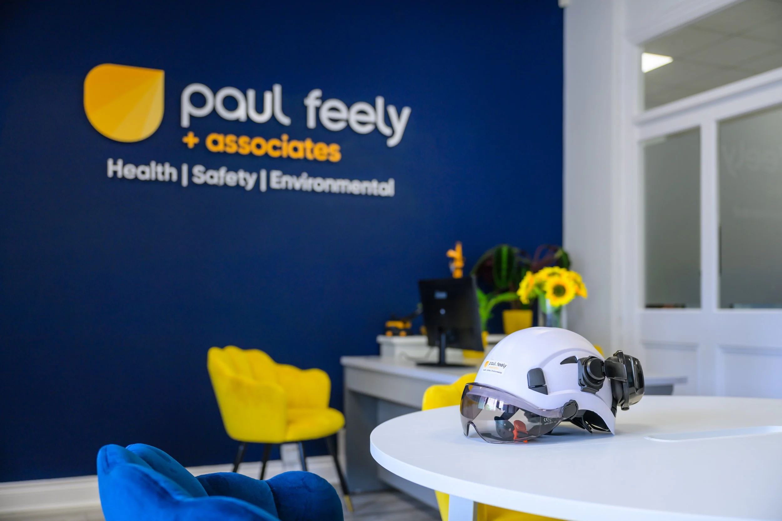 A white helmet with black goggles placed on a white table in a modern office or reception area. In the background, there is a dark blue wall with the logo and name of 'Paul Feely + associates' with the tagline 'Health | Safety | Environmental.' The workspace includes yellow and blue chairs, a computer on a white counter, and a vase with sunflowers and green leaves.