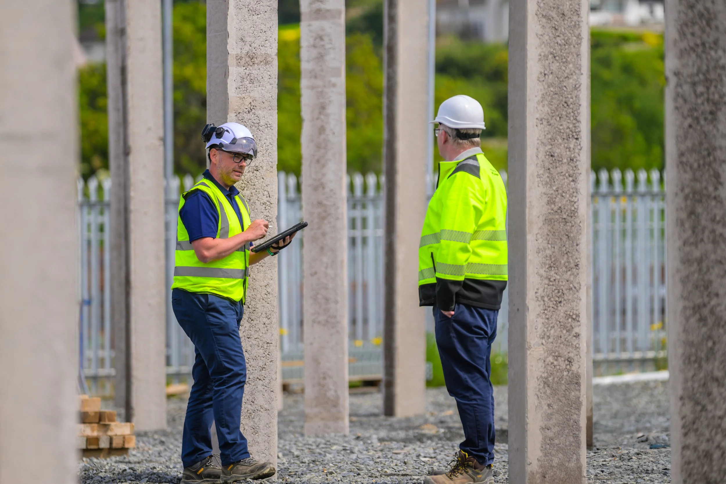 Two construction workers wearing safety helmets and high-visibility vests stand among concrete posts at a construction site on a cloudy day.