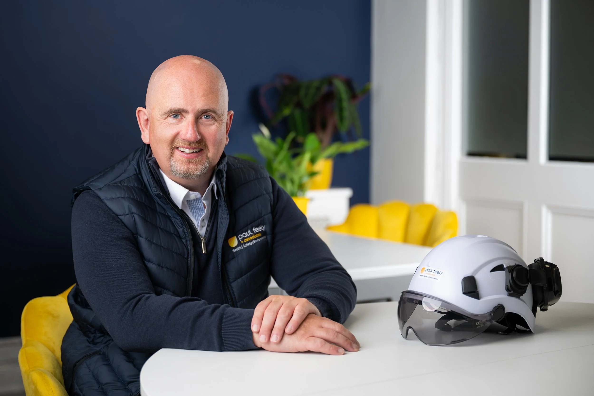 A middle-aged man with a shaved head and goatee, wearing a navy blue vest with a company logo, sitting at a white table with a white safety helmet and goggles, in a modern room with yellow chairs and plants.