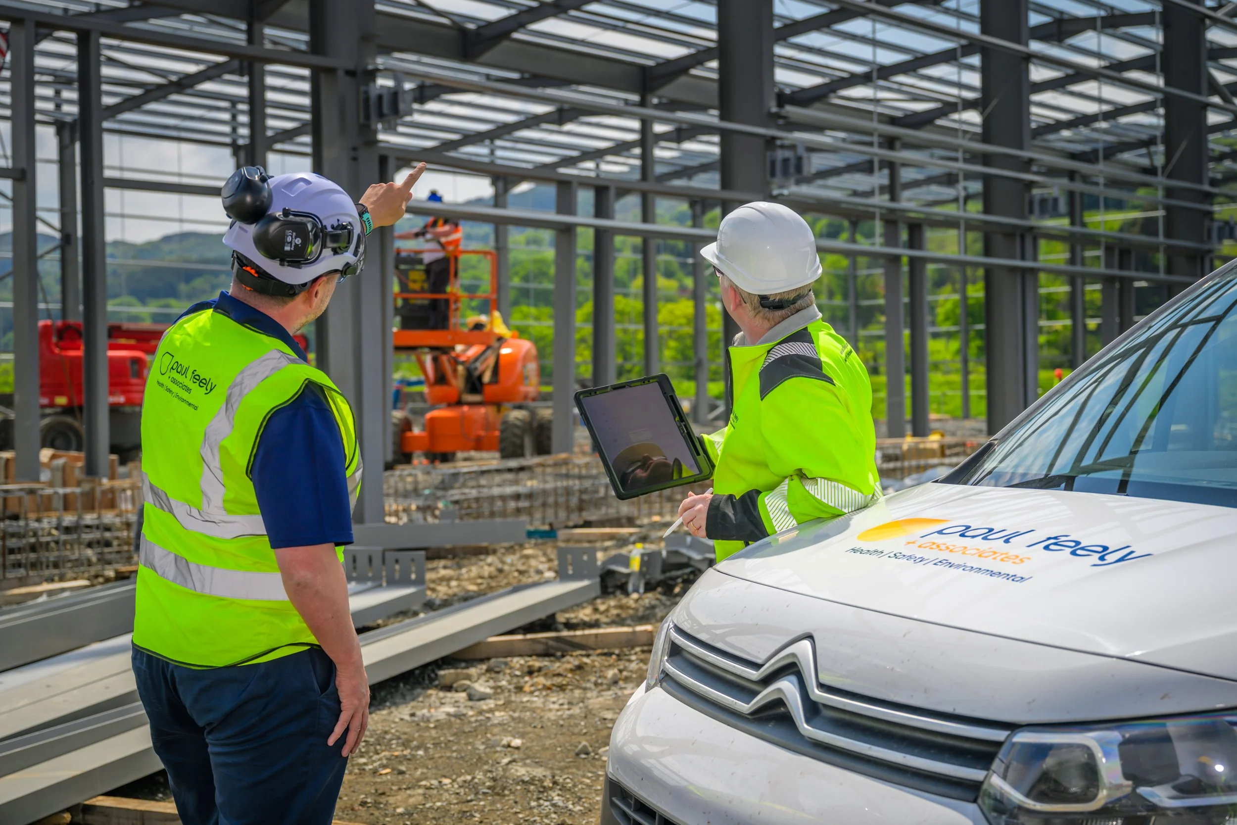 Two construction workers in high-visibility jackets and hard hats are having a discussion at a construction site with a partially built steel framework in the background. One worker is pointing, and the other holds a tablet. A white company vehicle with the logo 'Paul Feely' is parked nearby.