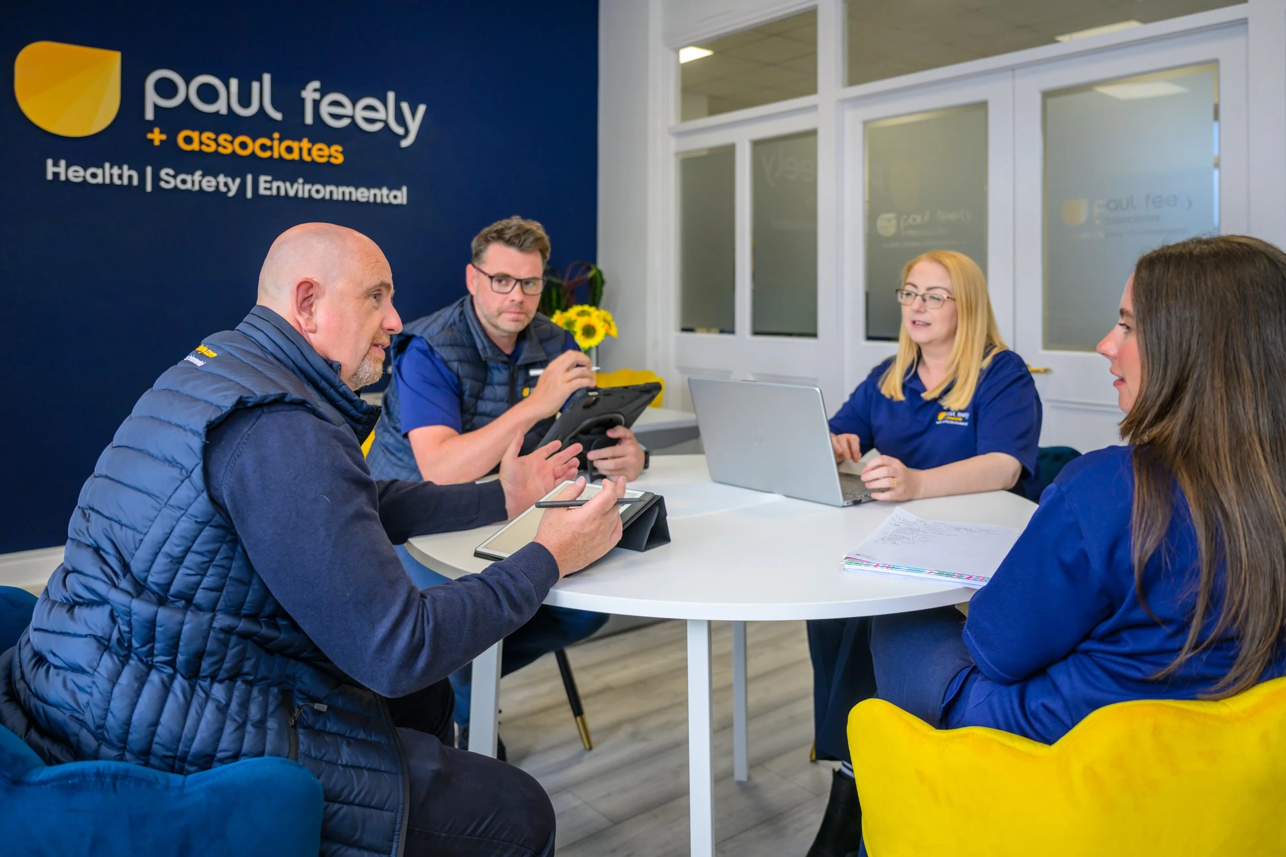 Four people sitting around a white table in a conference room, engaged in a discussion. There is a navy blue wall with the logo and branding for 'Paul Feely + associates' in the background. The room has glass windows and a bright, professional atmosphere.