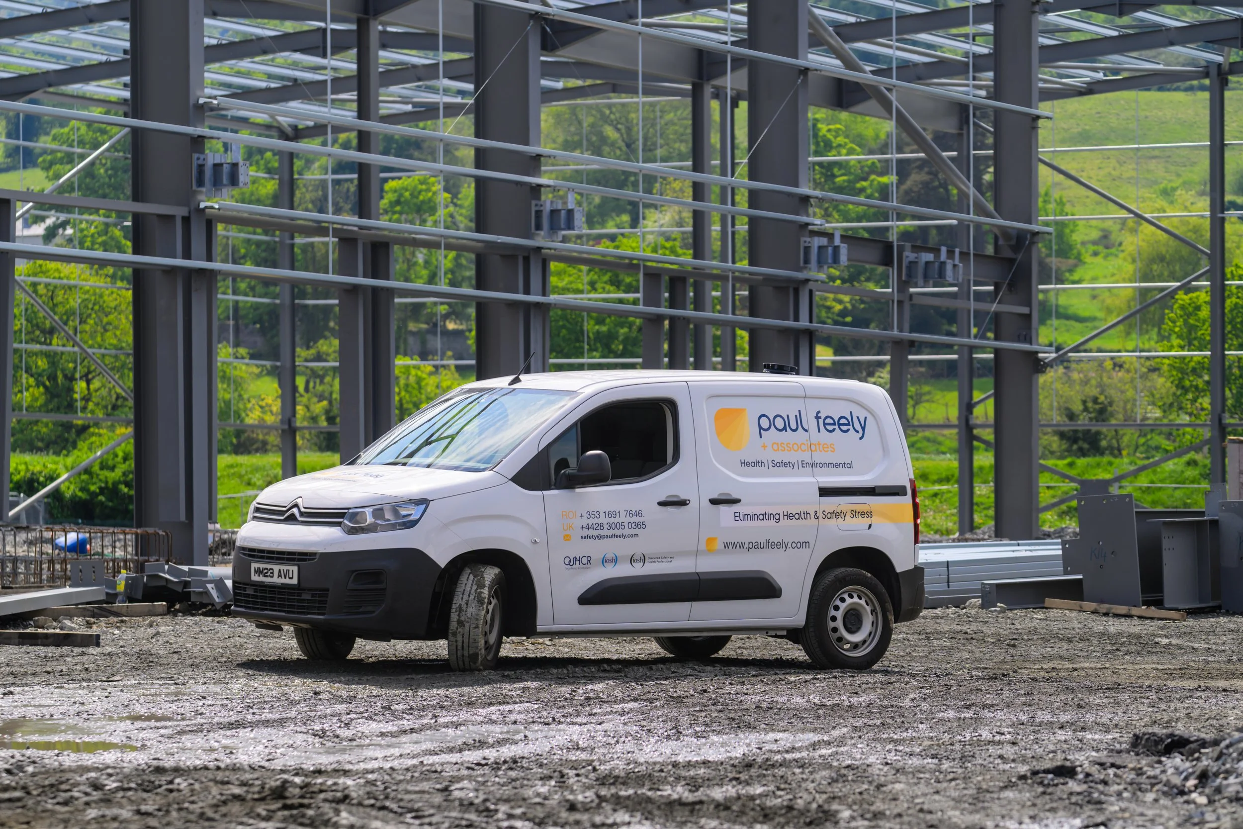 A white Paul Feely + Associates van parked on a construction site with metal framework in the background and green hills with trees.