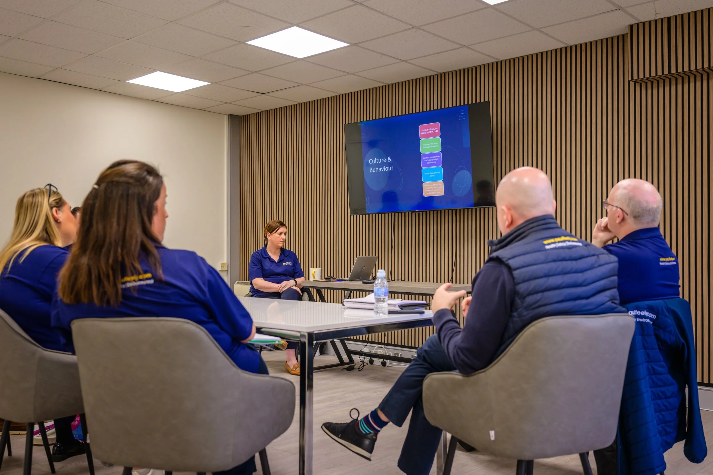 A group of people attending a presentation in a conference room. The presenter is a woman sitting at a desk with a laptop, facing a screen displaying a slide titled "Culture & Behaviour". The audience members are wearing matching uniforms, and the ro