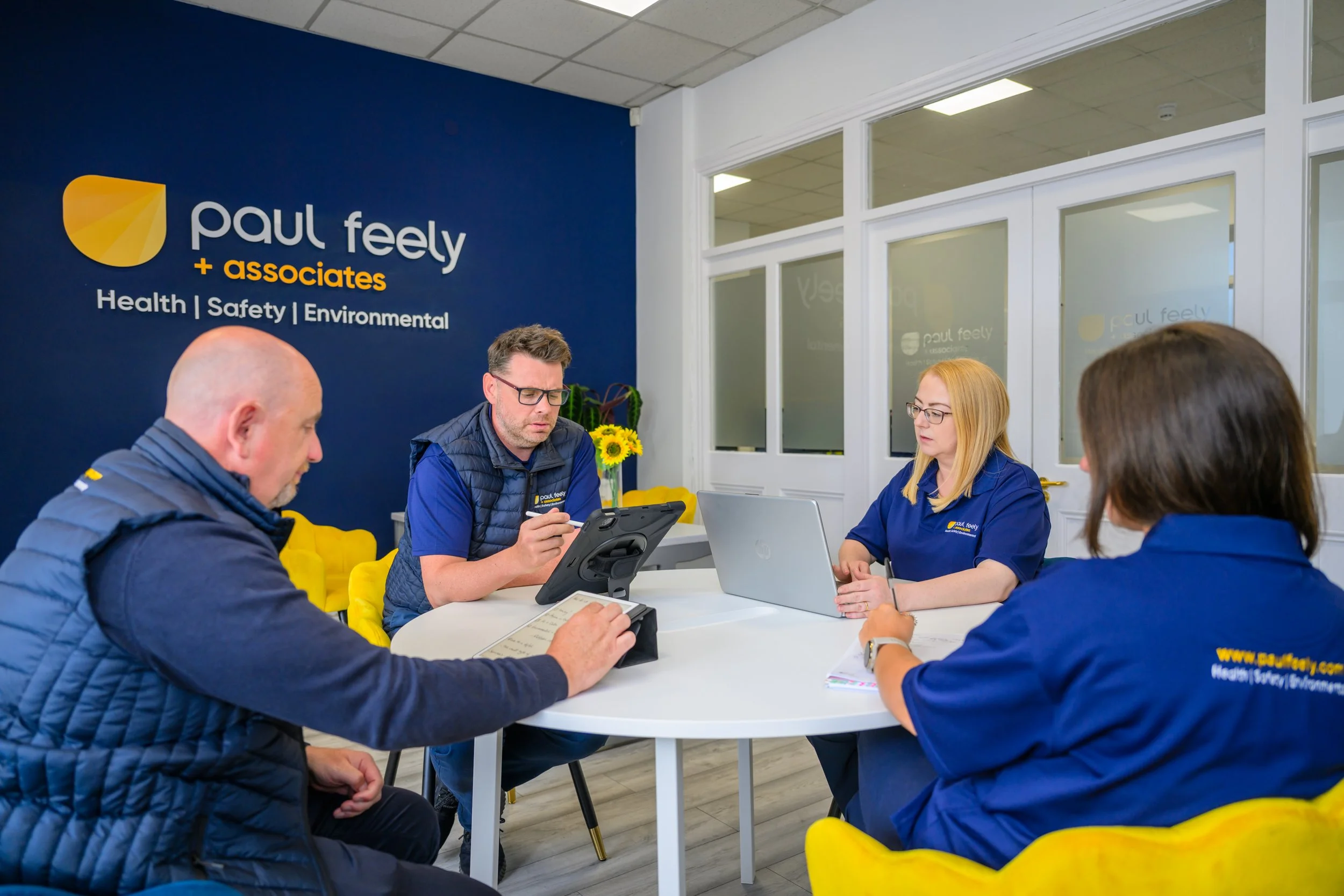 Four people sitting around a white table in a meeting room at Paul Feely & Associates, with a blue wall displaying the company's name and services for health, safety, and environmental consulting; two women and two men, with laptops, tablets, and not