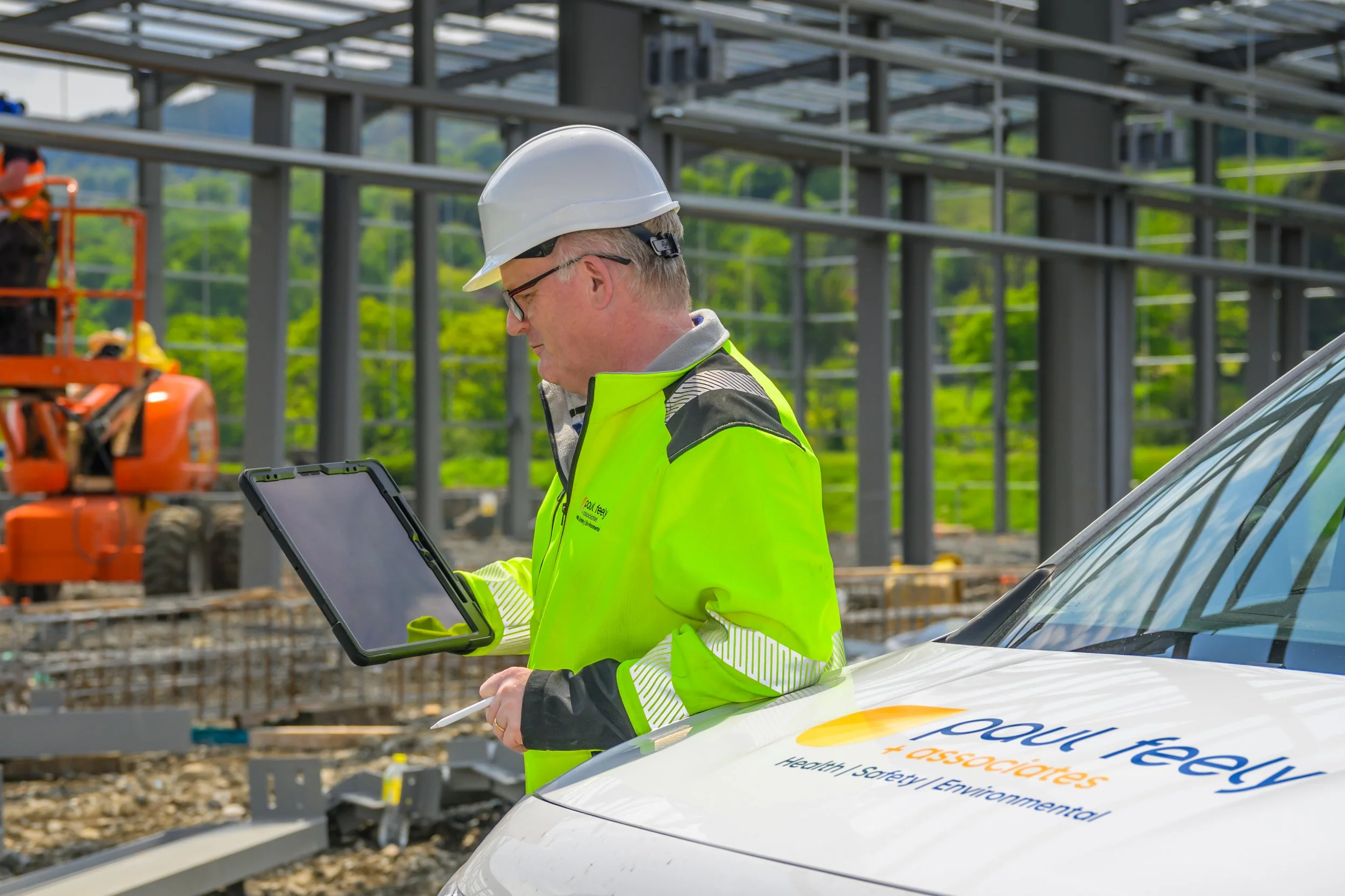 A man wearing a white hard hat and a high-visibility yellow jacket with reflective stripes is standing next to a white vehicle at a construction site. He is looking at a tablet device and appears to be inspecting or reviewing information. In the background, there is construction equipment and a structure with metal beams, with greenery visible behind the site.